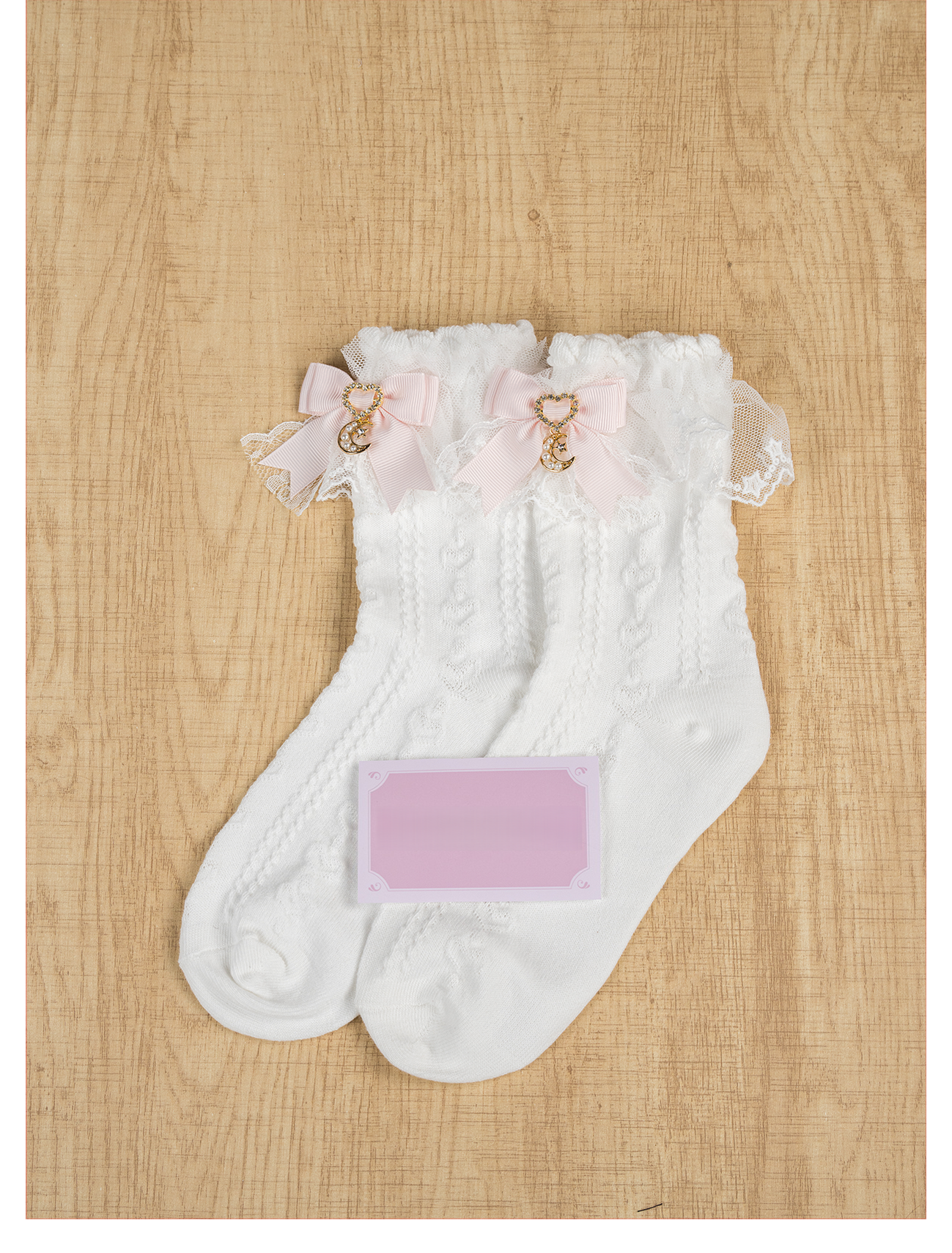 Light Pink Jirai Kei ankle socks with ruffled lace cuff and pink bow moon charm pendant, flat lay on wooden surface