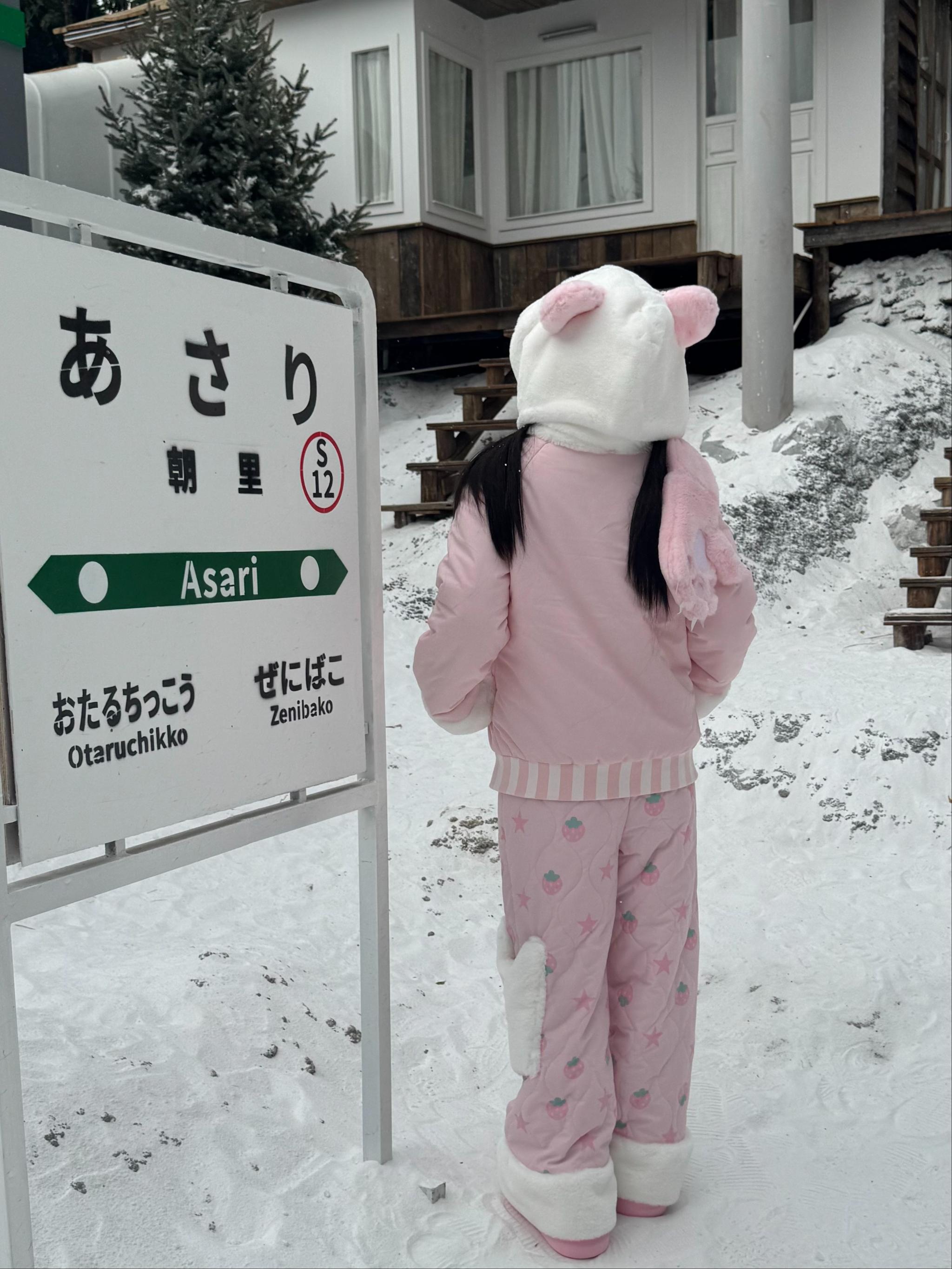Back full-body view of pink Jirai Kei quilted cotton winter outfit featuring fuzzy white ankle cuffs subtle all-over fruit prints matching pink coat and kawaii aesthetic at snowy Japanese train stop