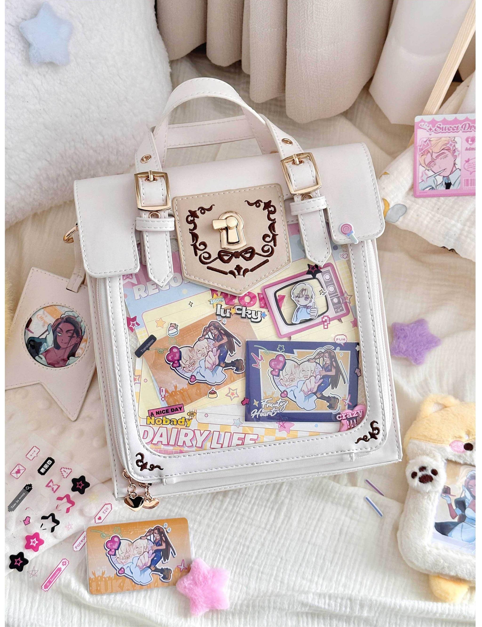 White vintage Jirai Kei ita satchel bag with gold metal lock hardware and clear front window packed with colorful anime stickers and collectible badges, flat lay indoor bedding scene