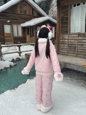 Back view of strawberry Jirai Kei pink winter padded coat with quilted pattern pants, white fluffy trim, on model at snowy riverside wooden cabin area