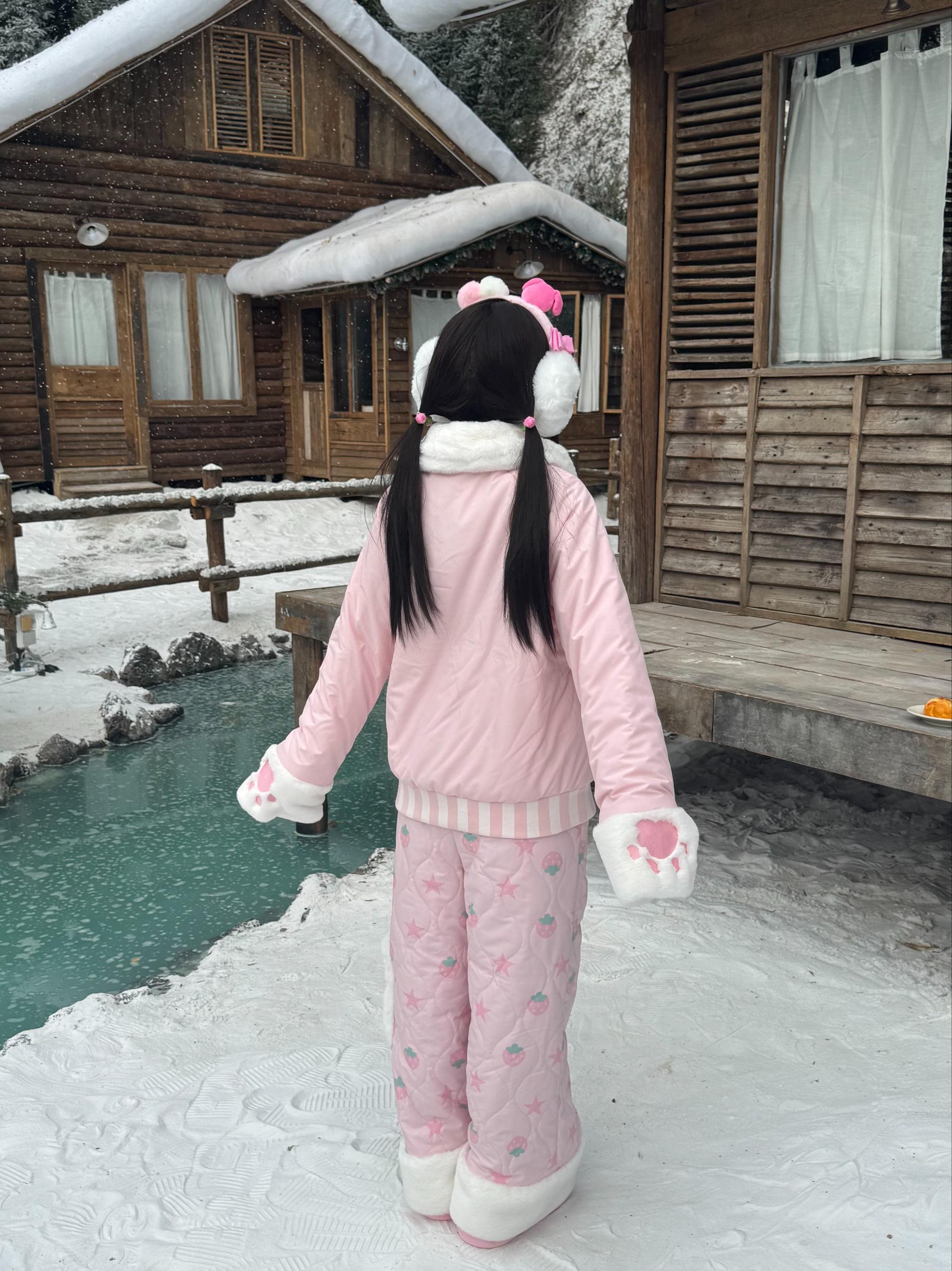 Back view of strawberry Jirai Kei pink winter padded coat with quilted pattern pants, white fluffy trim, on model at snowy riverside wooden cabin area