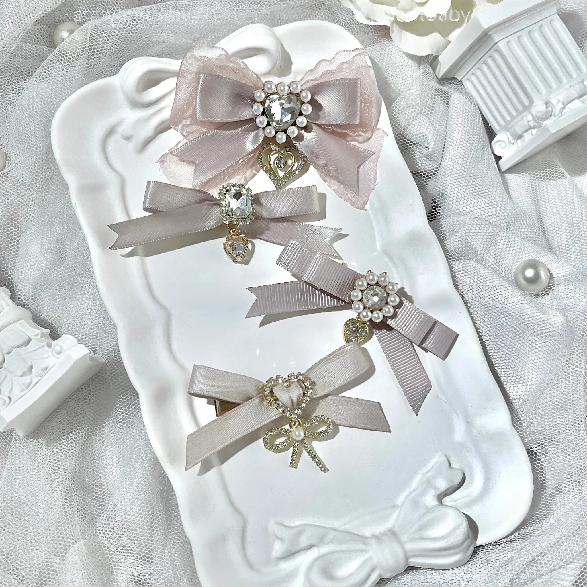 Beige cream mesh and satin bow hair clips set with rhinestone centers and heart pendants on decorative tray lace background top-down flat lay