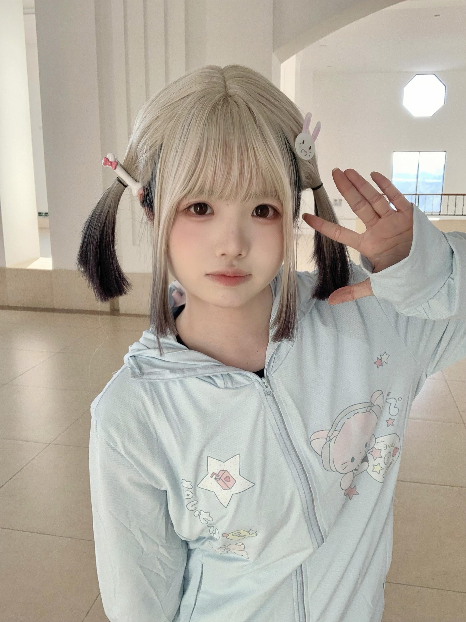 Front eye-level shot of kawaii jirai kei UV jacket in water blue with adorable cat and star graphics, modeled by blonde twin-tail girl in bright lobby