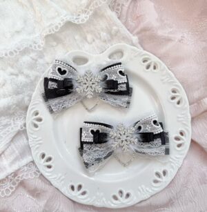 Jirai Kei black and white bow hair clips with lace cut-outs and silver snowflake charm on white decorative plate over pink lace fabric flat lay styling