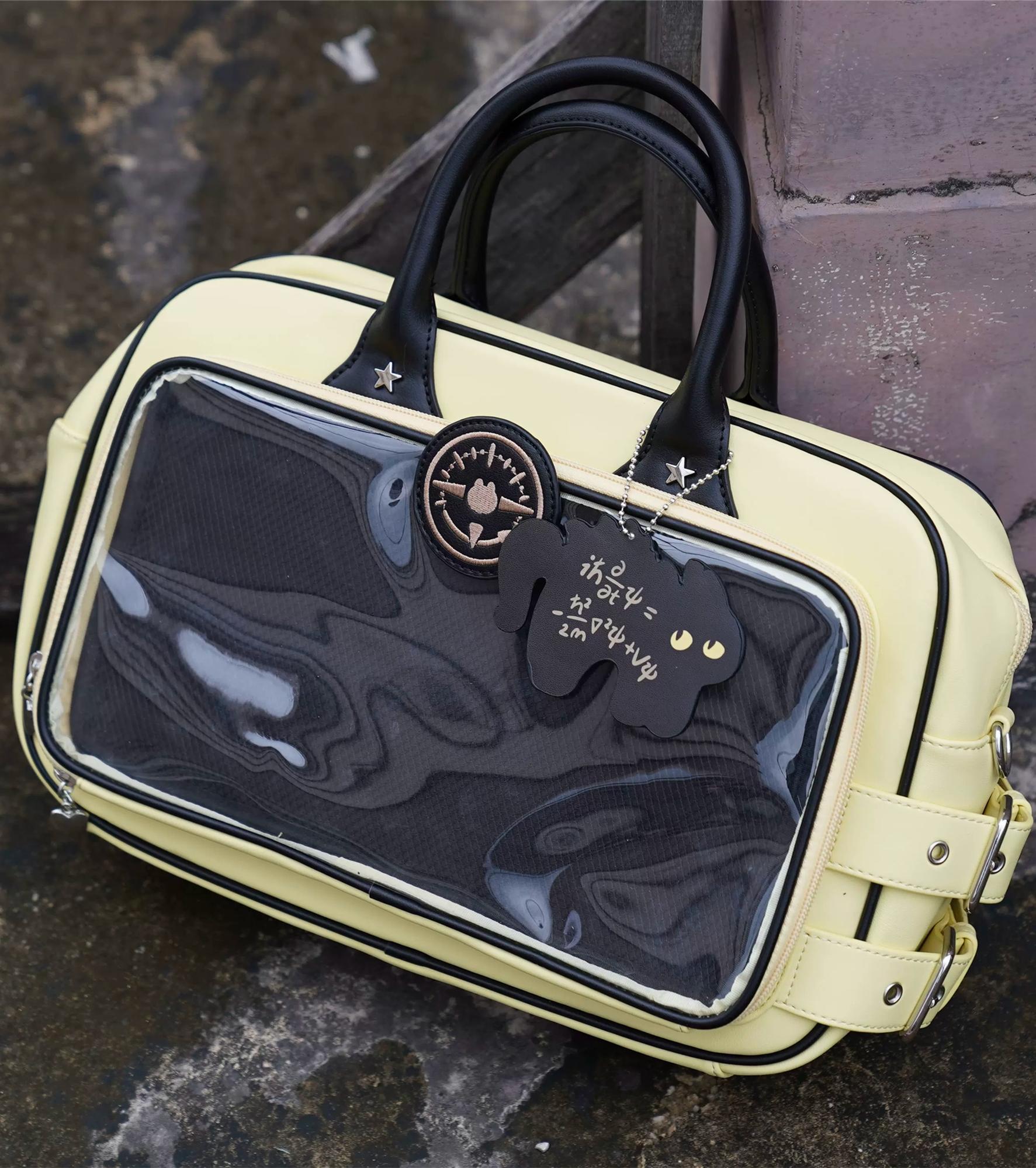 Cream and black Jirai Kei ita bag with large transparent front window featuring cat charm detail, close-up front view on outdoor stone surface backdrop