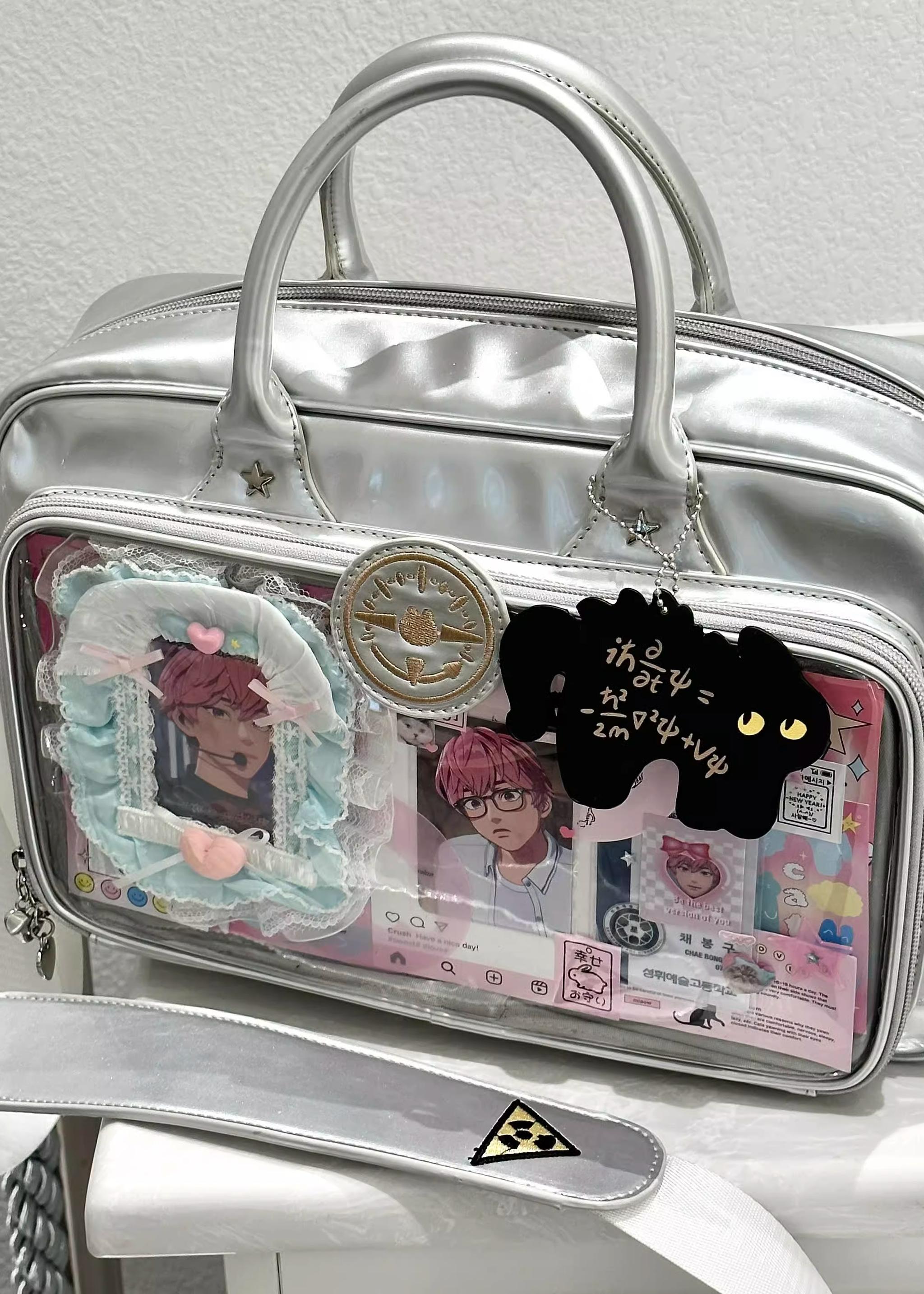 Silver metallic Jirai Kei ita shoulder bag with cat-shaped charm and anime badge decorations, close-up flat lay on studio desktop showing shine and detail
