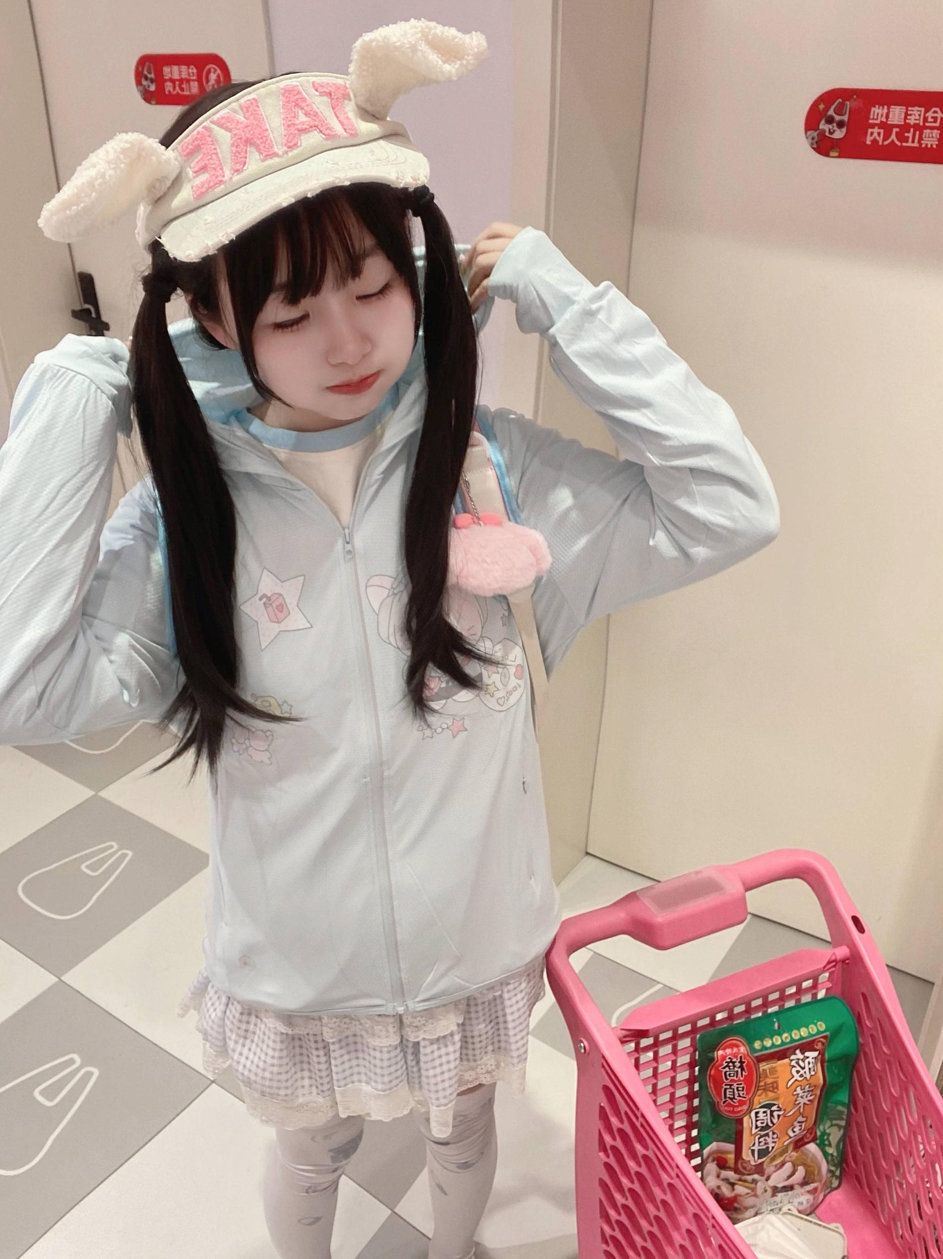 Full-body view of jirai kei sun jacket on dark twin-tail girl wearing bunny ear hat, standing near pink shopping cart in convenience store aesthetic