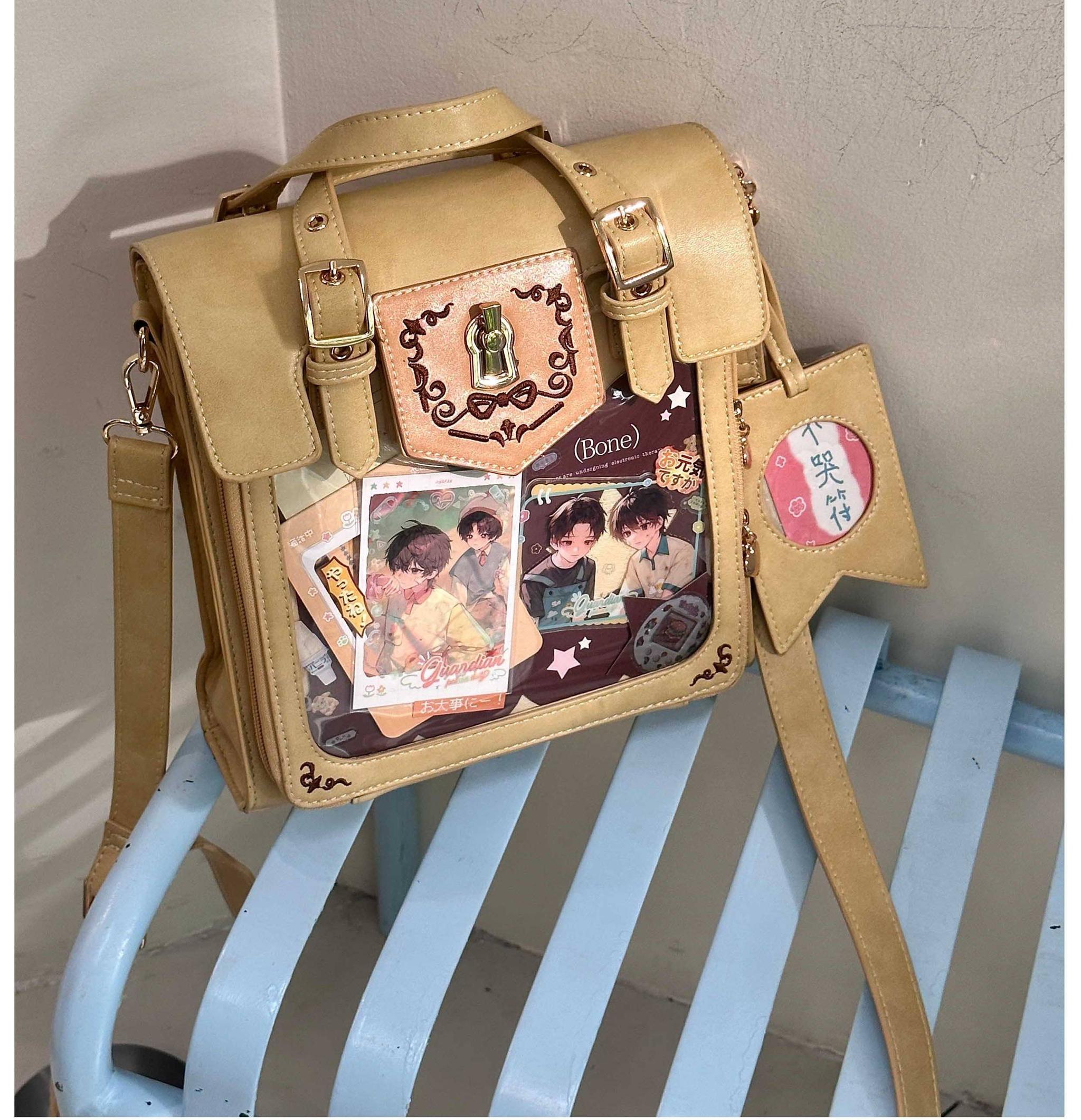 Khaki vintage Jirai Kei ita satchel bag with detachable shoulder strap and gold decorative hardware, clear window with anime prints visible, three-quarter angle outdoor bench scene