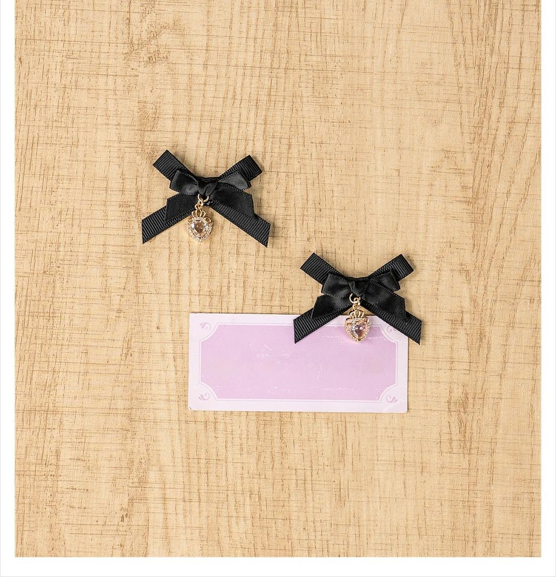 Pair of black mini bow hair clips with dangling heart-shaped metal charm on wooden table flat lay