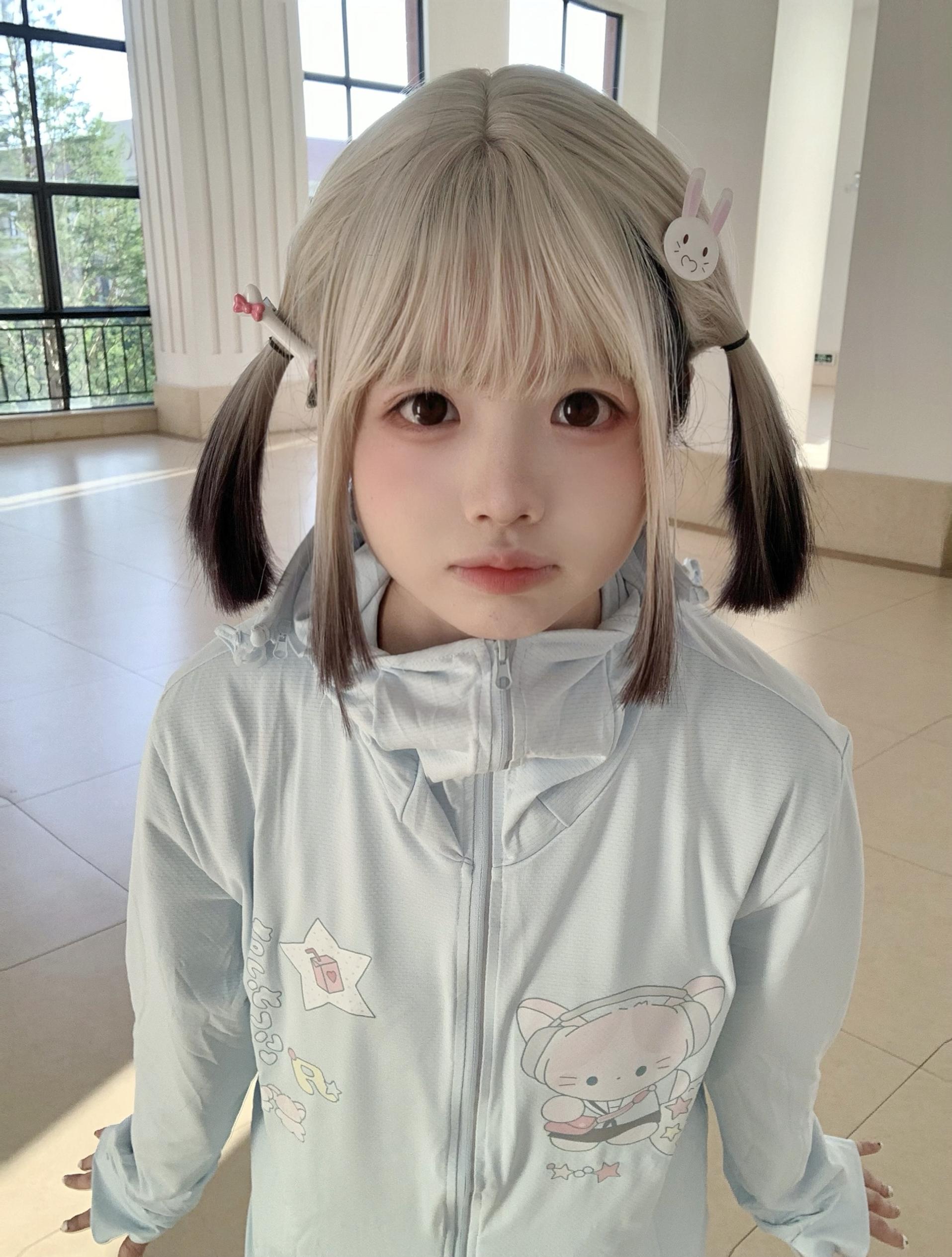 Front view of kawaii jirai kei water blue UV protection jacket with cute cat and star prints, modeled by blonde girl with twin-tails, indoor lobby setting with natural light