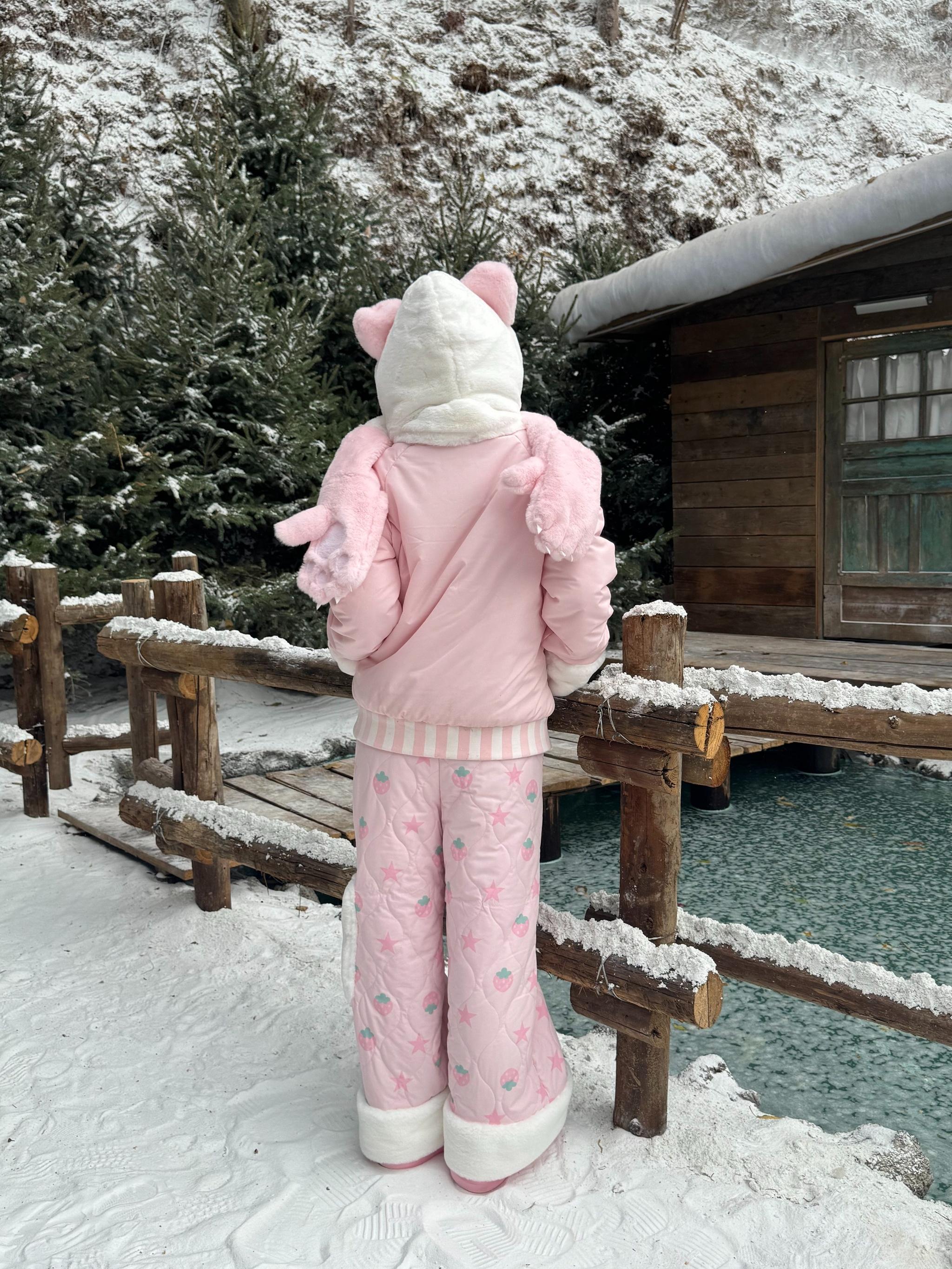 Full back view of strawberry Jirai Kei quilted winter cotton set showing bear ear hood plush wing decorations on jacket fuzzy white trims and all-over print by snowy lakeside wooden cabin