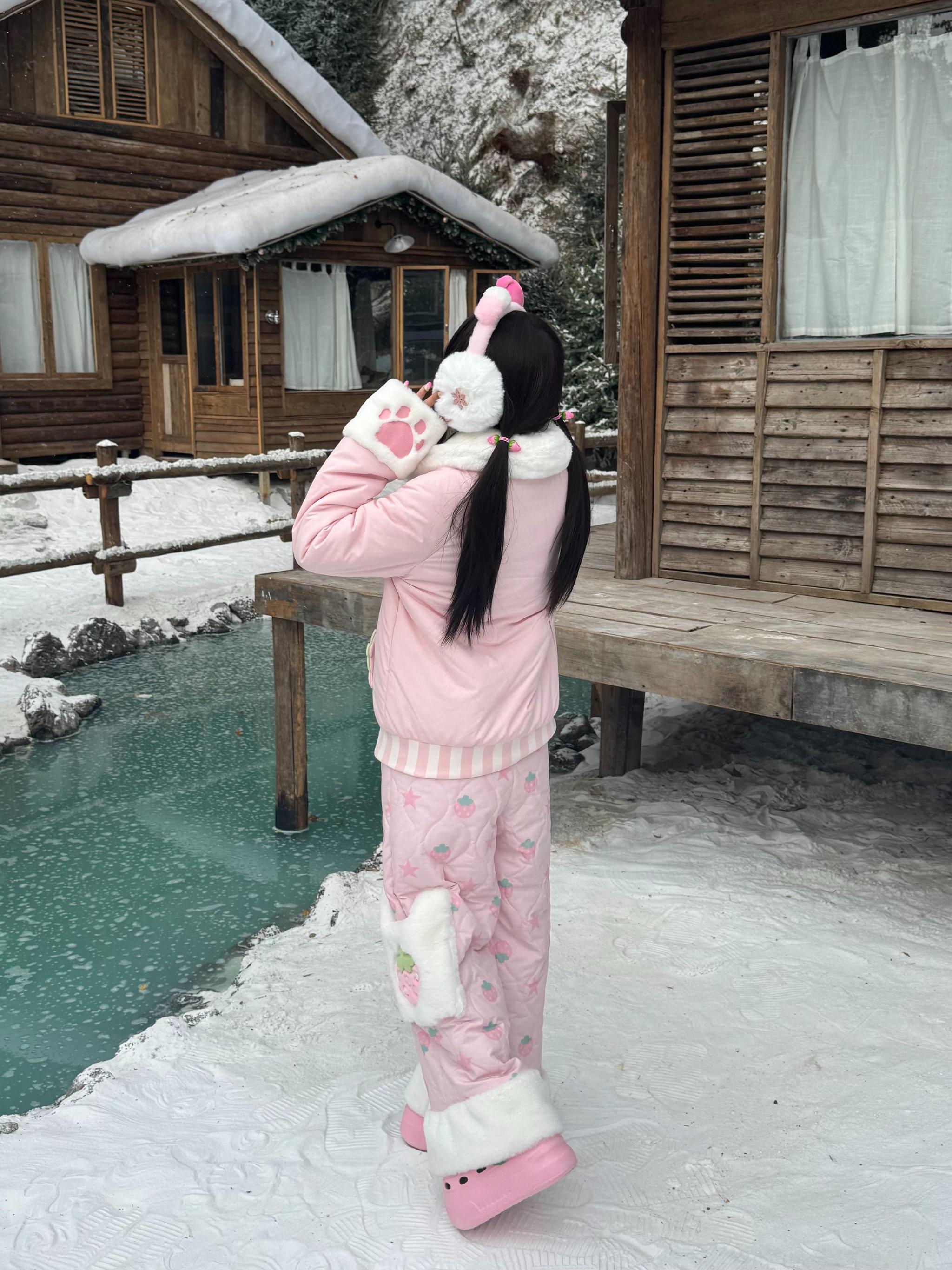 Back three-quarter view of pink Jirai Kei padded winter coat with bow-adorned ear warmers, pink shoes, strawberry pattern pants at snowy riverside cabin deck