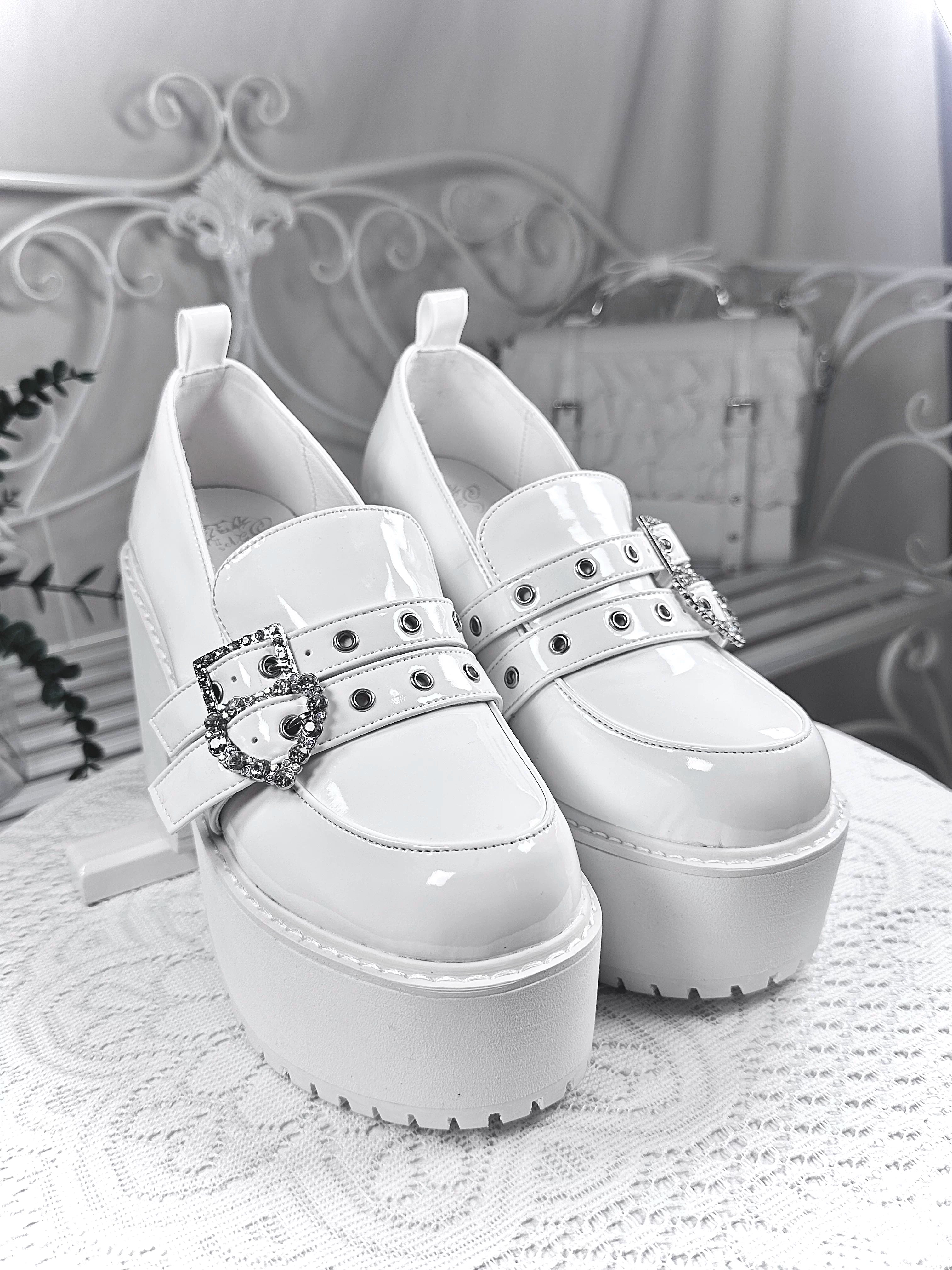 White patent leather platform loafers front overhead view on lace tabletop showcasing studded straps thick platform sole and heart rhinestone buckles detail