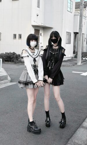 Pair of masked female models in Jirai Kei outfits featuring white off-shoulder sailor collar top with black gothic styling ensemble, demonstrating versatile layering options with sailor blouse as core piece
