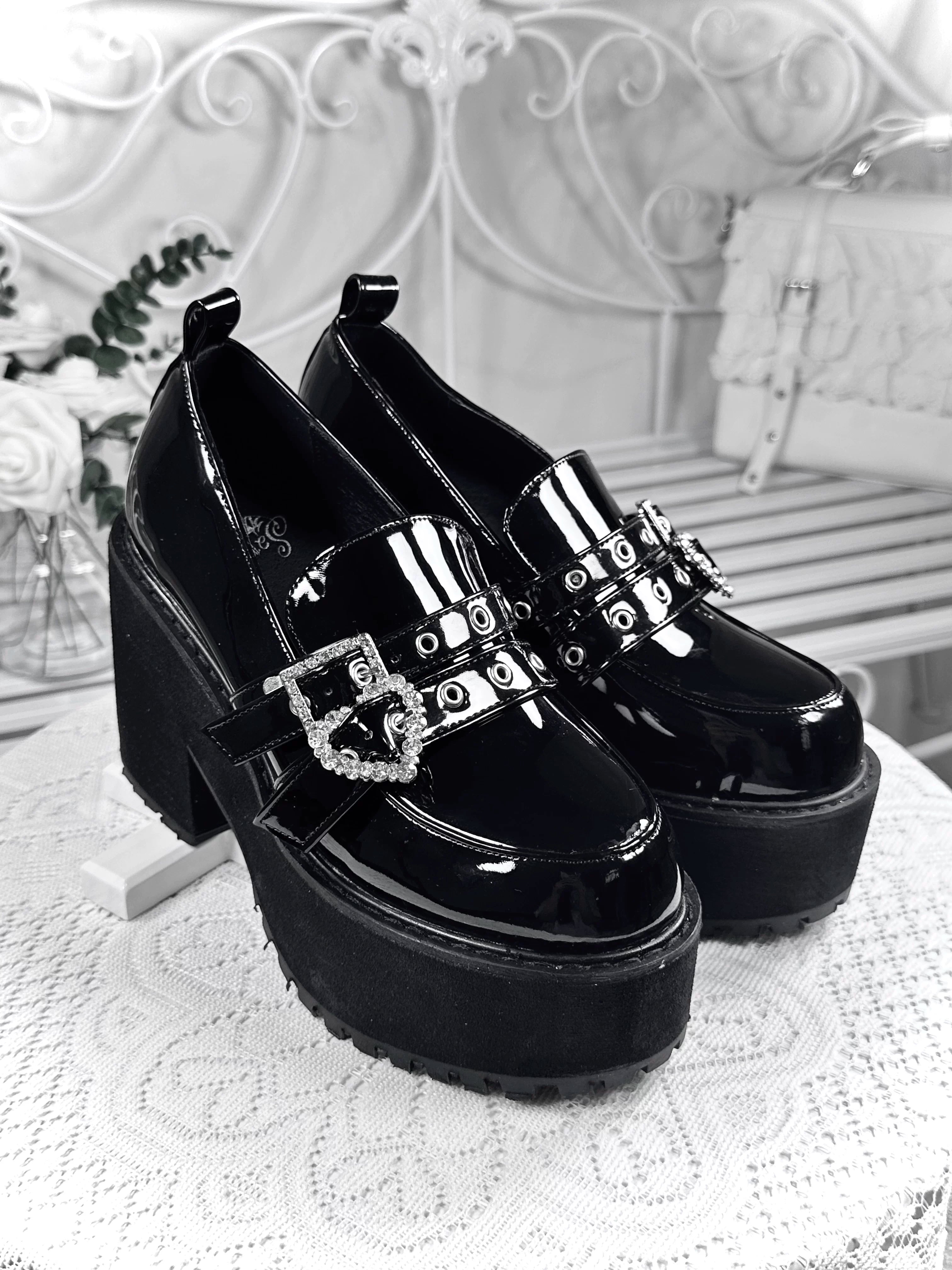 Black patent leather platform loafers three-quarter overhead view on lace tabletop with chunky block heel heart rhinestone buckles in vintage bedroom setting