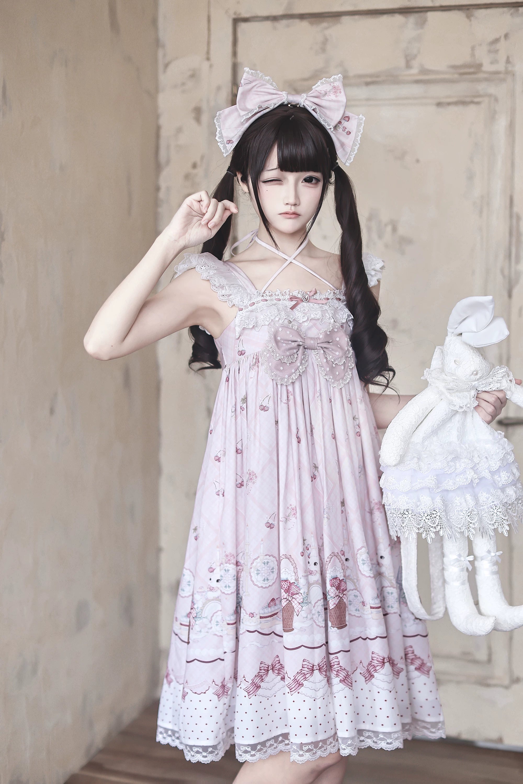 Close-up front medium shot of strawberry bunny Jirai Kei Lolita dress featuring cross halter neck front chest bow with model holding lace rabbit doll accessory
