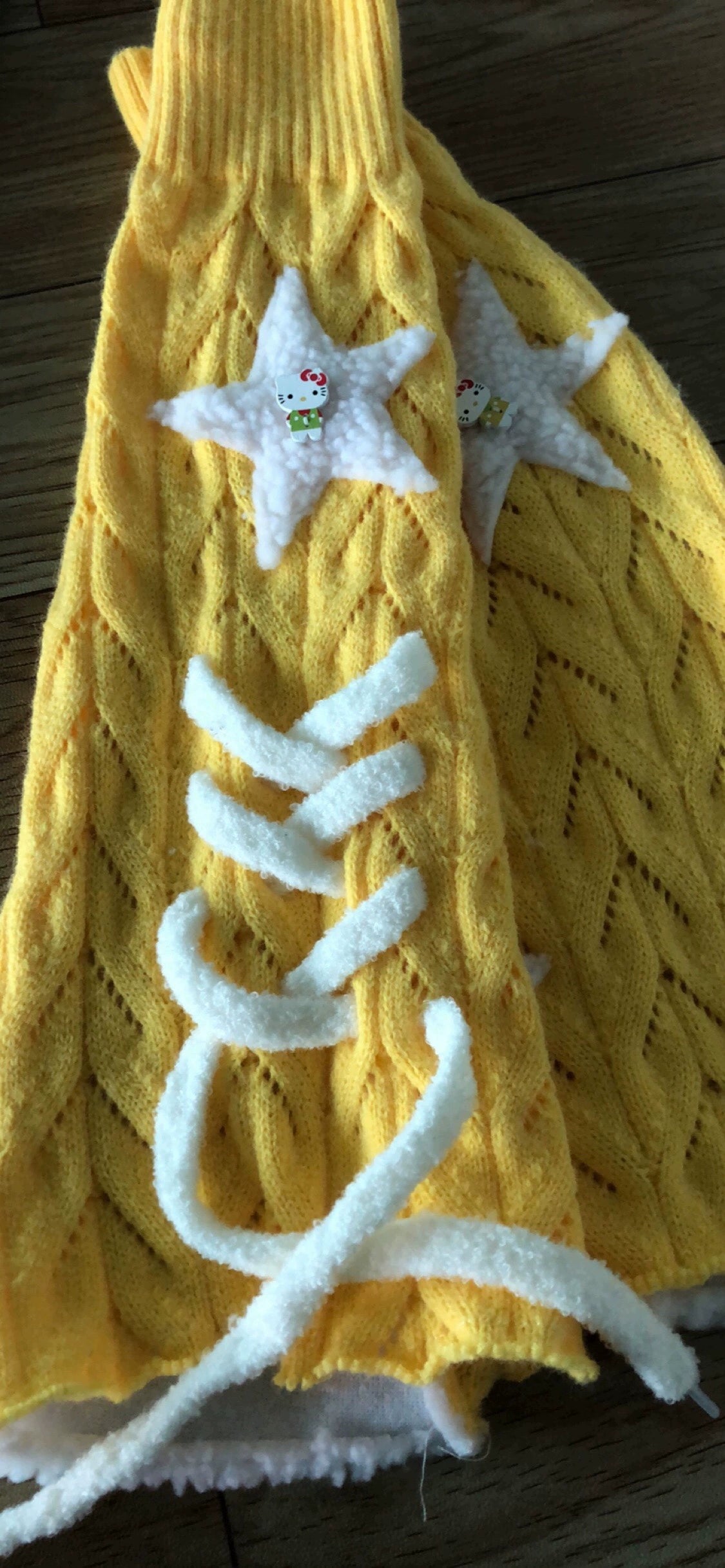 Flat lay close-up of sunny yellow Jirai Kei leg warmers on wooden desktop, featuring hollow knit construction with white lace-up straps and fluffy star decorations, artfully frayed hem edges detail