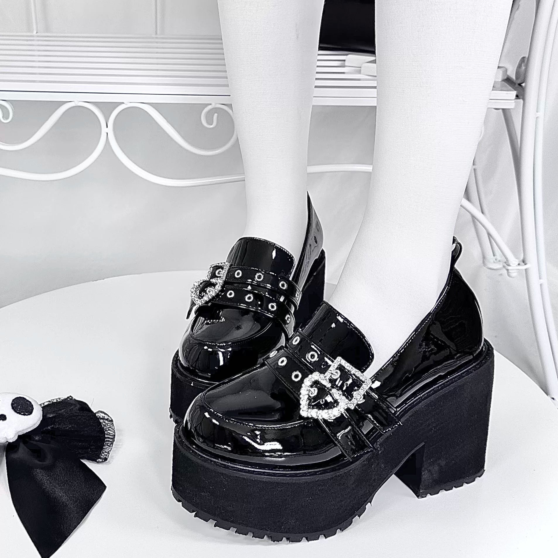 Black patent leather platform loafers frontal eye-level view on model with white stockings featuring chunky block heel and heart-shaped rhinestone buckles