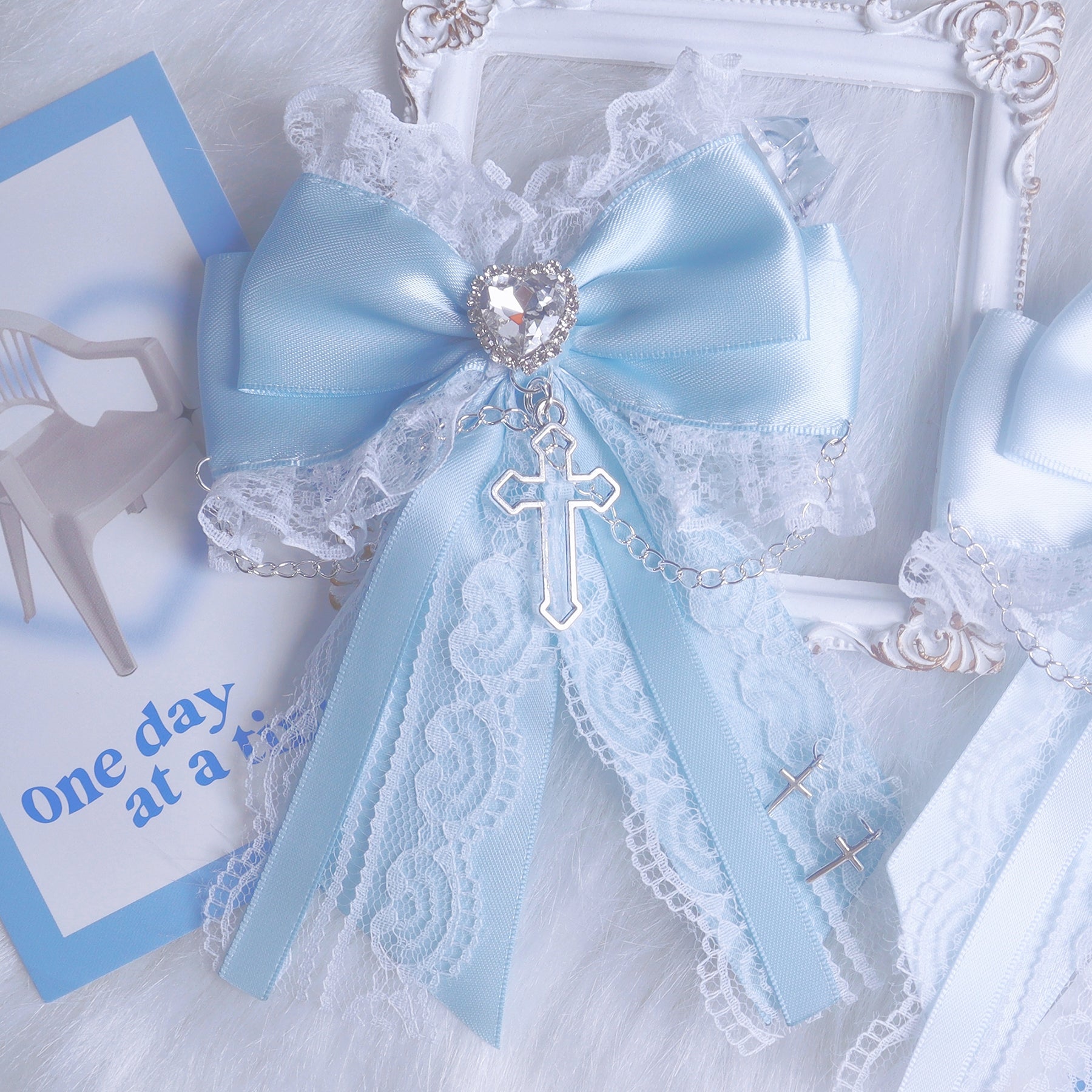 Light blue satin bow hair clip side view close-up on stationery background showing lace edge heart-shaped rhinestone and cross pendant details