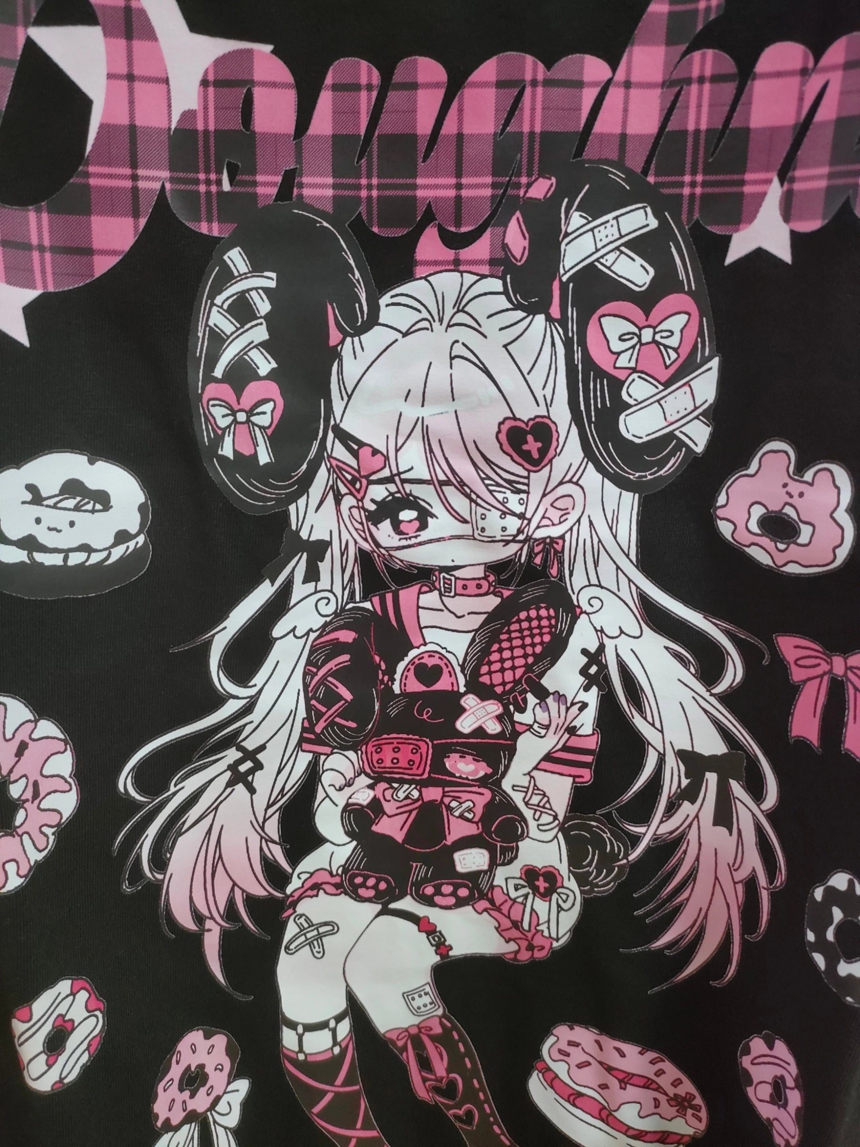 Flat lay front view of pink and black Jirai Kei anime T-shirt print featuring pink anime girl graphic, pink plaid lettering, pink black white color scheme with donut decorations