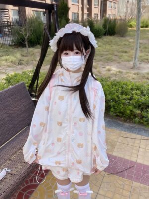 Twin-tailed girl wearing pastel bear print Jirai Kei sunscreen jacket with cat ears hood and matching shorts, outdoor residential setting