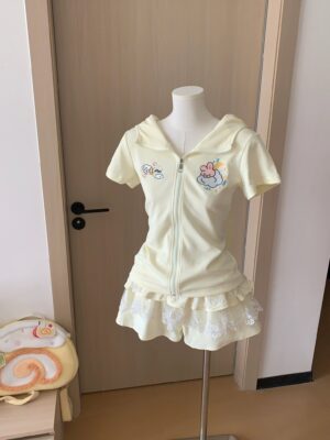 Front view of bunny ear jirai kei yellow zip-up hoodie with adorable cartoon embroidery and short sleeves paired with pale yellow tiered ruffles lace skirt, displayed on mannequin with door background