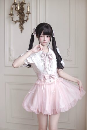 Front eye-level portrait of soft pink ryousangata tulle bubble hem skirt with rhinestone waist bow and white bow-decorated blouse against elegant chandelier wall backdrop - jirai kei lolita fashion