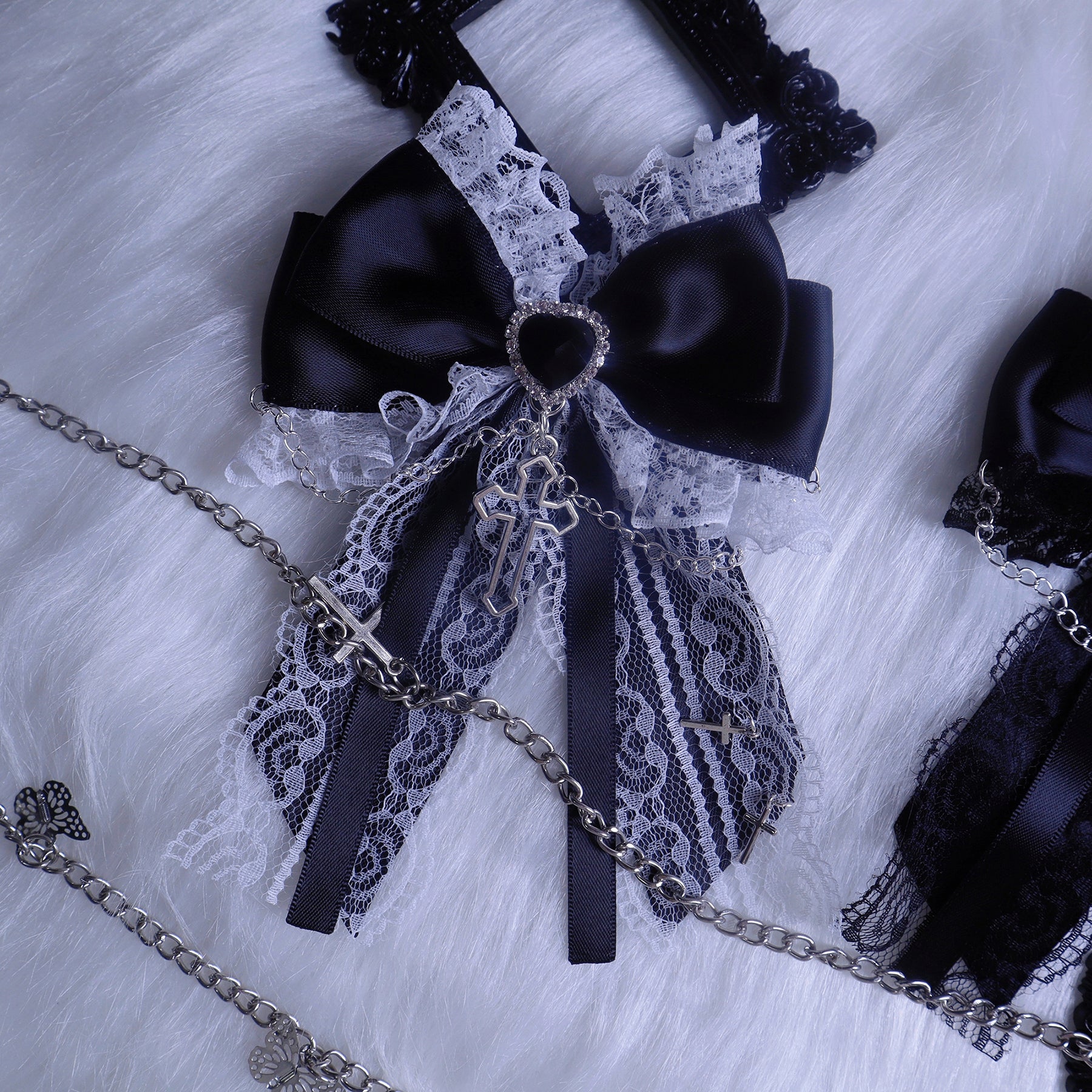 Black-navy dual-tone bow hair clip close-up flat lay on fuzzy fabric displaying lace overlay, heart-shaped center stone and silver cross chain pendant