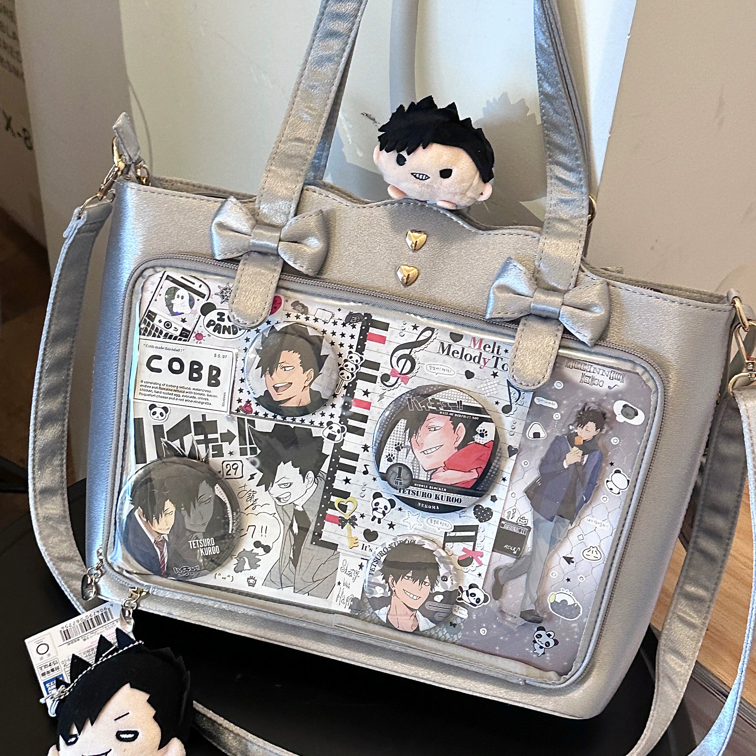Silver gray Jirai Kei ita bag front view on tabletop with adorable plush doll decoration on top, bow embellishments, clear transparent badge display panel, and removable adjustable shoulder strap