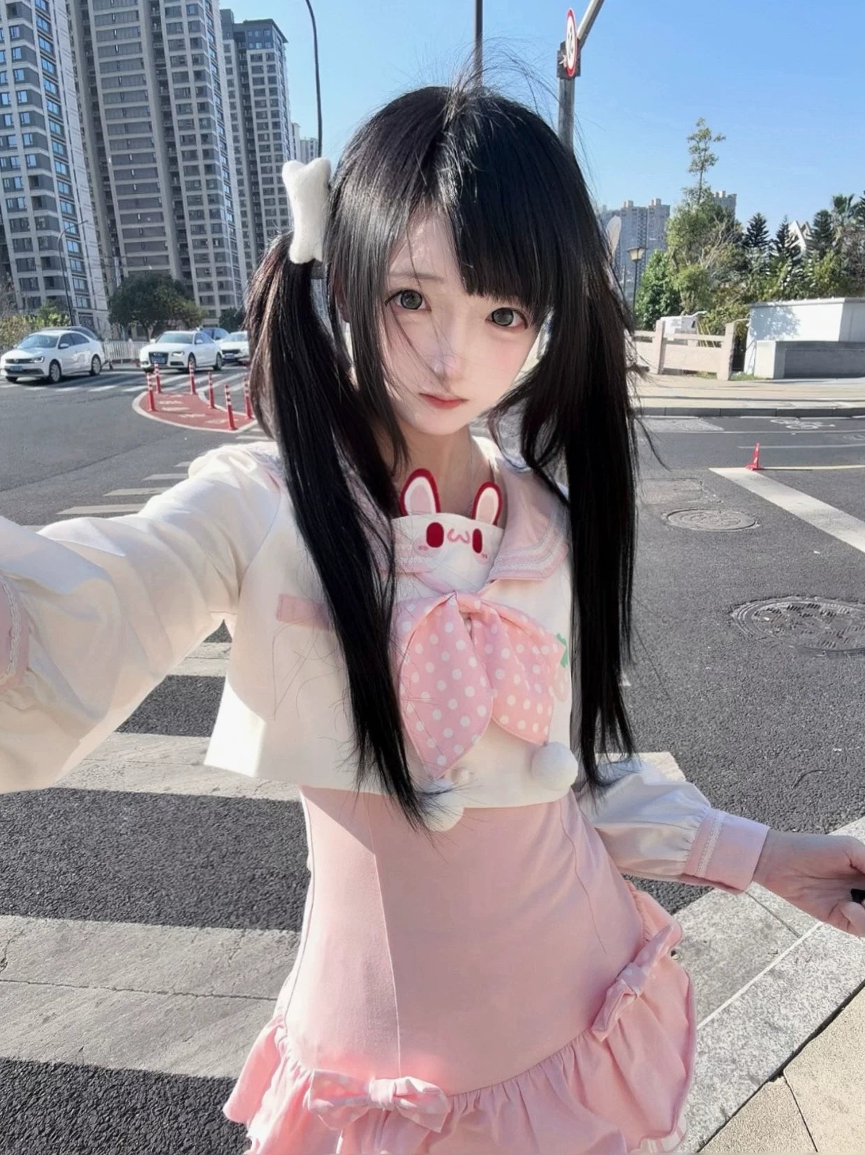 Close-up selfie angle of kawaii sailor collar Jirai Kei blouse with rabbit chest ornament, polka-dot pink removable bow collar, ruffle mini skirt on urban street