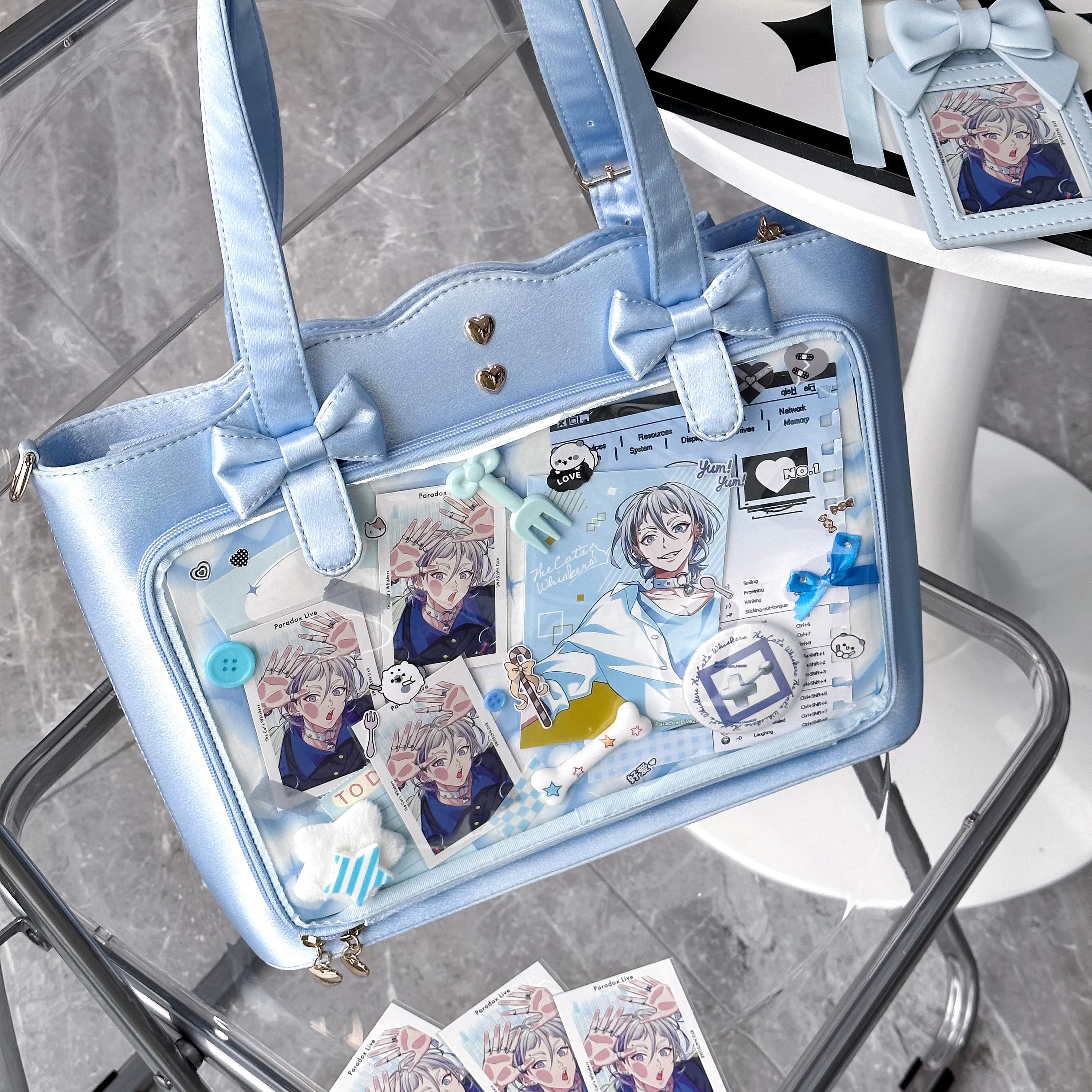 Sky blue Jirai Kei ita bag overhead tabletop view next to white table, displaying heart metal studs, bow decorations, clear anime character pin display panel, and removable adjustable crossbody strap