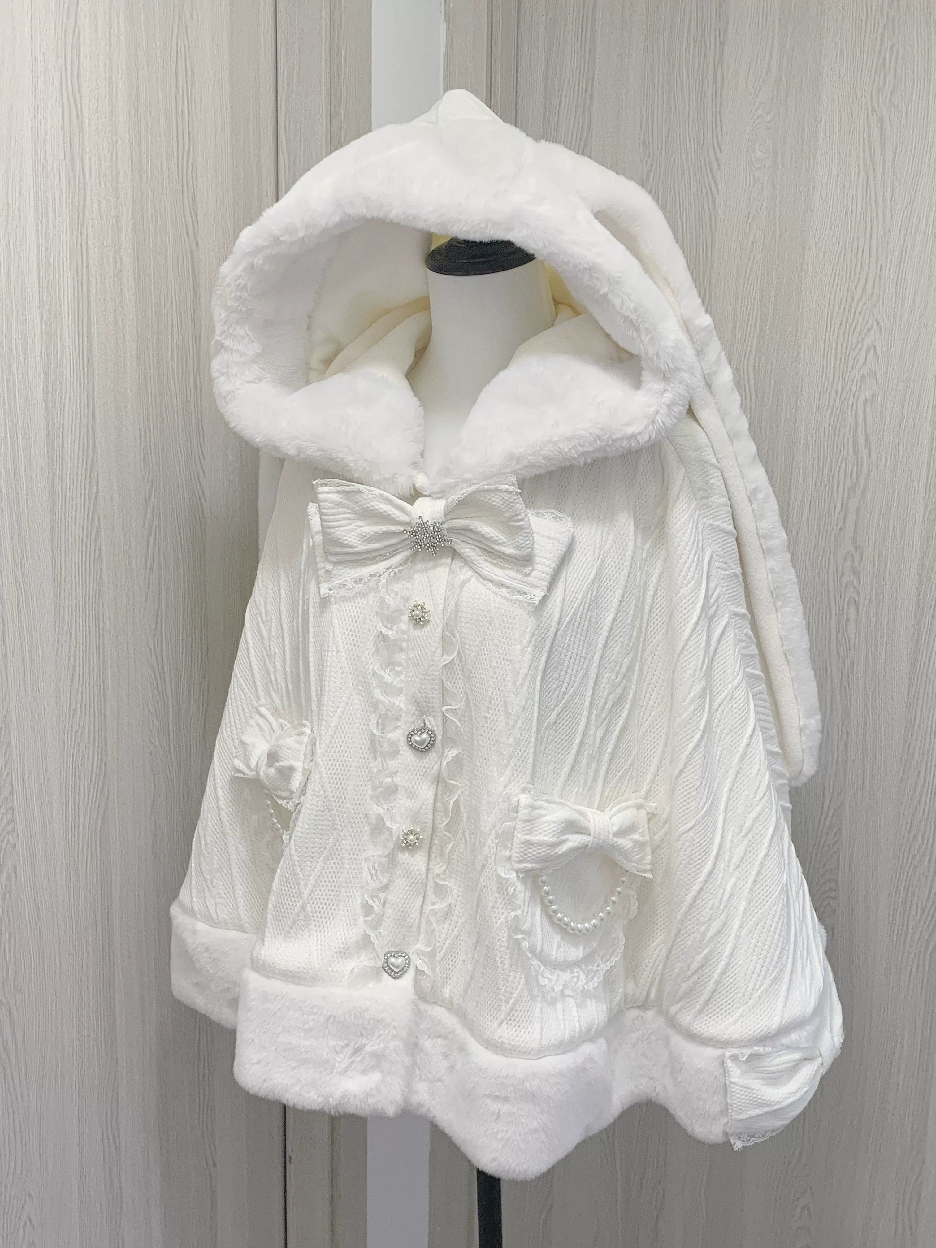 Static front display view of white rabbit ear fleece cape coat on full body mannequin, showing rabbit ear hood, front bow decoration, button closure, and pocket bows
