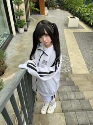 Black and white striped Jirai Kei sports jacket outdoor styling on model with twin-tails hairstyle, showing full front view of jacket with chest embroidered patch and contrasting sleeve stripe details in natural courtyard setting