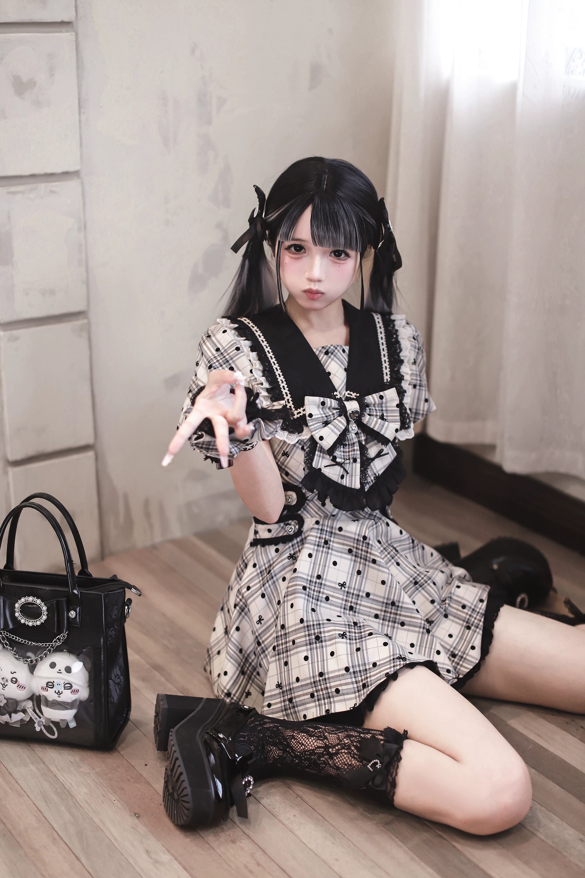 Medium kneeling shot of model wearing black lace socks and platform shoes with plaid polka dot Jirai Kei dress, showcasing skull-print handbag accessory in vintage room