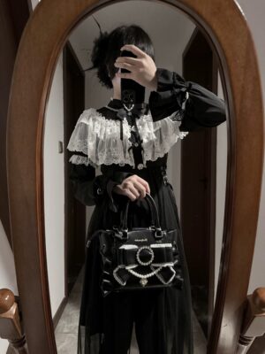 Black Jirai Kei handbag worn by gothic lolita model in partial mirror selfie view showing rhinestone pearl bow ornament, ruffled leather edges, and handbag styling