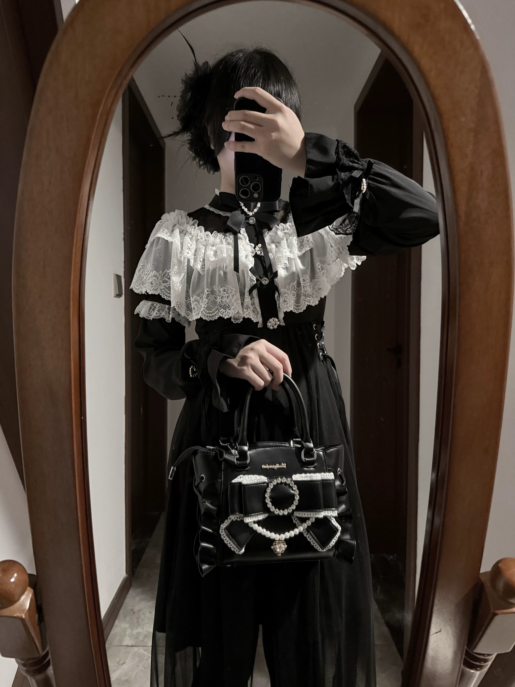 Black Jirai Kei handbag worn by gothic lolita model in partial mirror selfie view showing rhinestone pearl bow ornament, ruffled leather edges, and handbag styling
