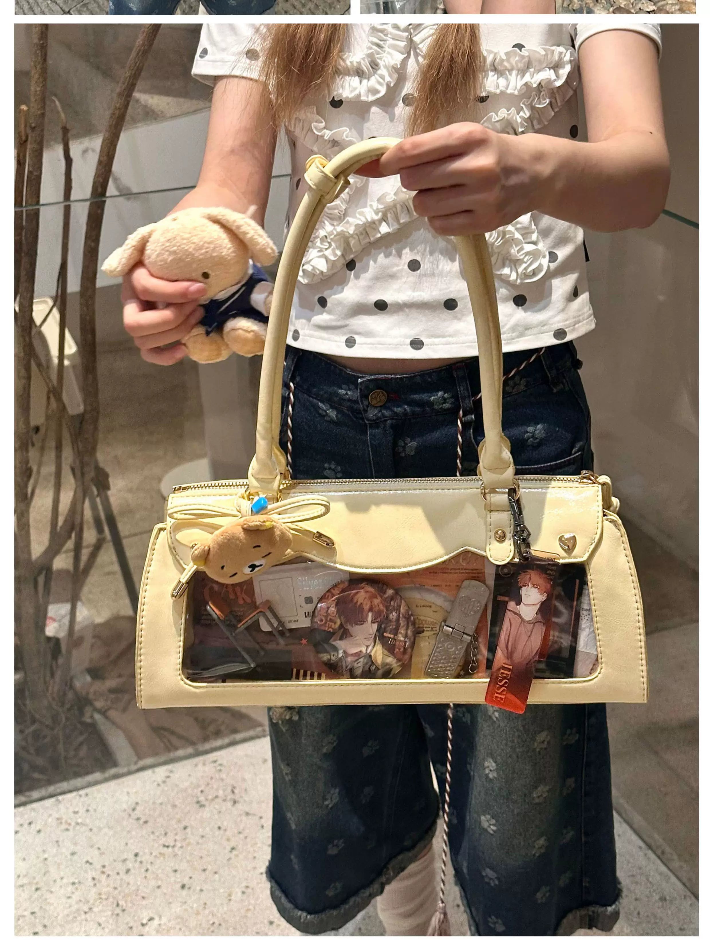 Cream and beige baguette itabag with small plush charm and clear display panel, outdoor street front angle view