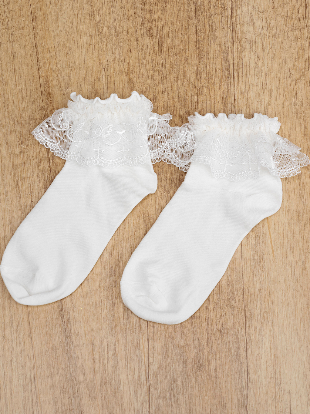Pair of white Jirai Kei lace ruffle ankle socks flat lay on wood surface sheer embroidered lace trim cuffs design showcase