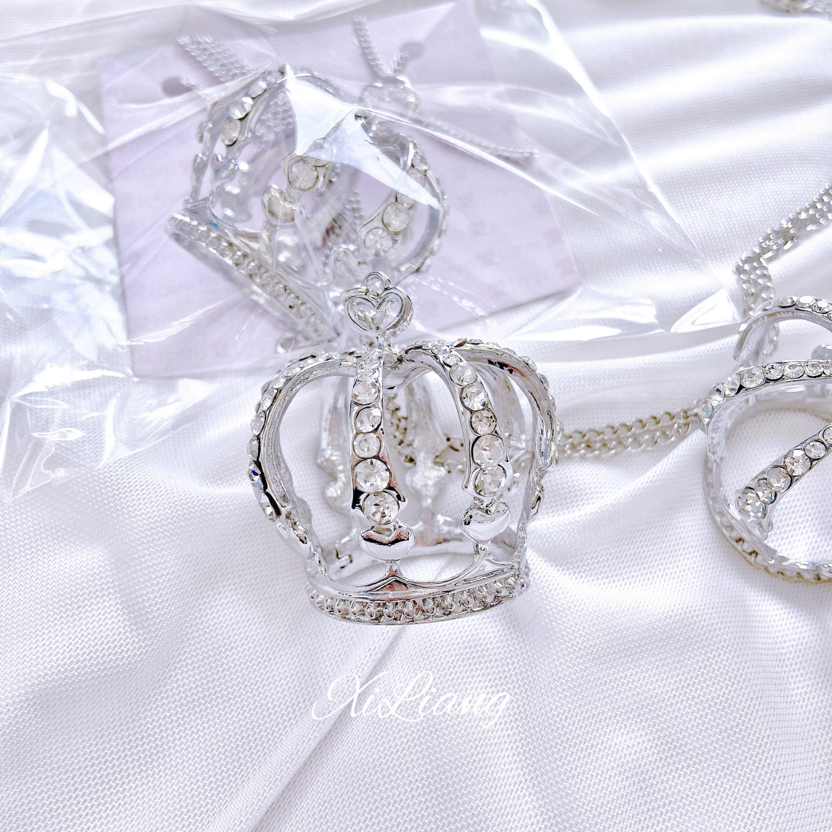 Large crown rhinestone necklace close-up on white satin fabric, silver hollow crown with heart-shaped chain connector fully covered with sparkling rhinestones detail shot