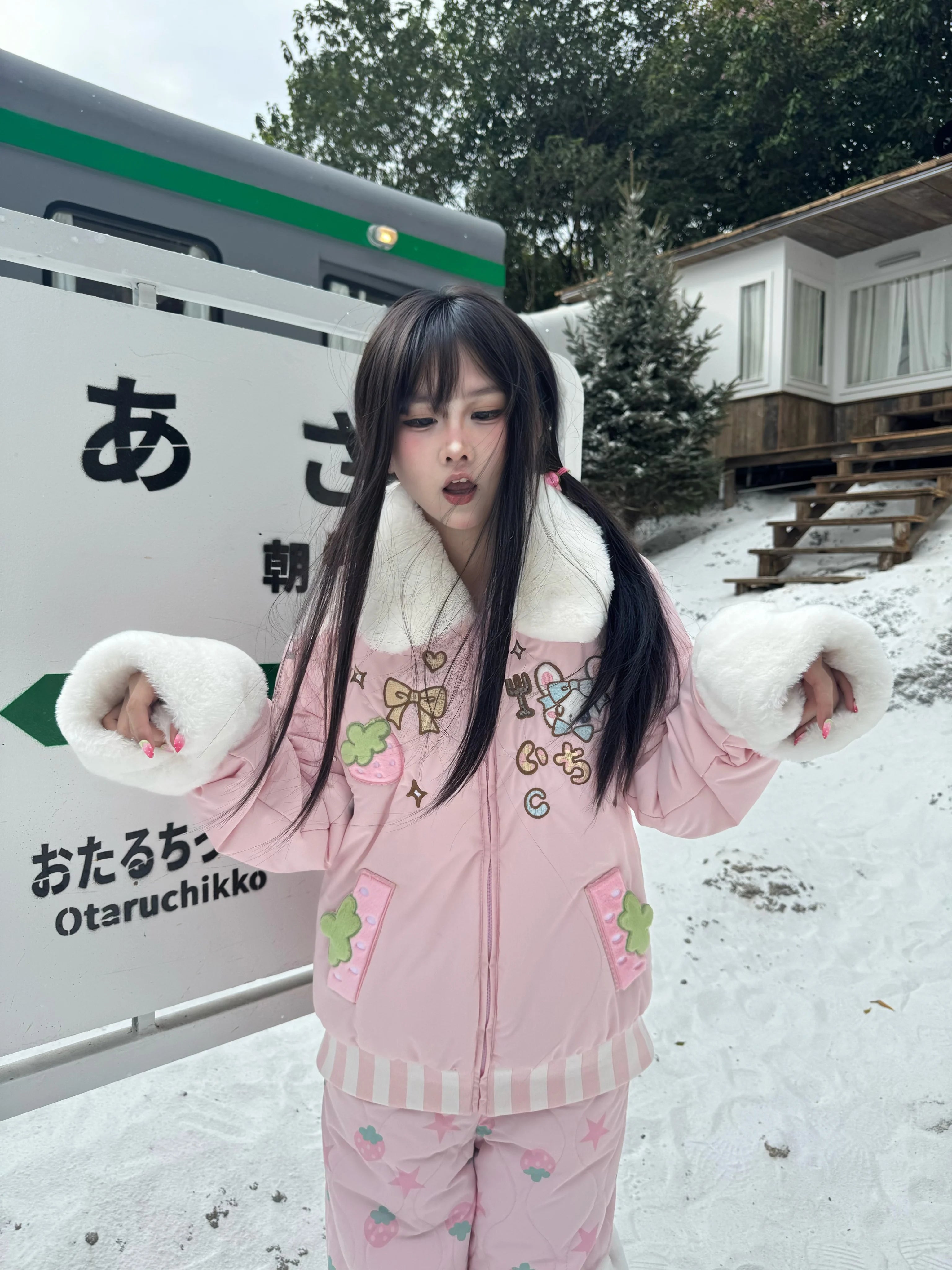 Close-up front detail of pink Jirai Kei padded coat with cartoon embroidery, white fuzzy trim, and strawberry motif pants at Japanese snowy train station