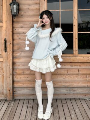 Light blue one-shoulder Jirai Kei sweater with furry neck trim and pom-pom bell sleeves paired with white layered tiered ruffle skirt, full body front standing view on outdoor wooden cabin deck