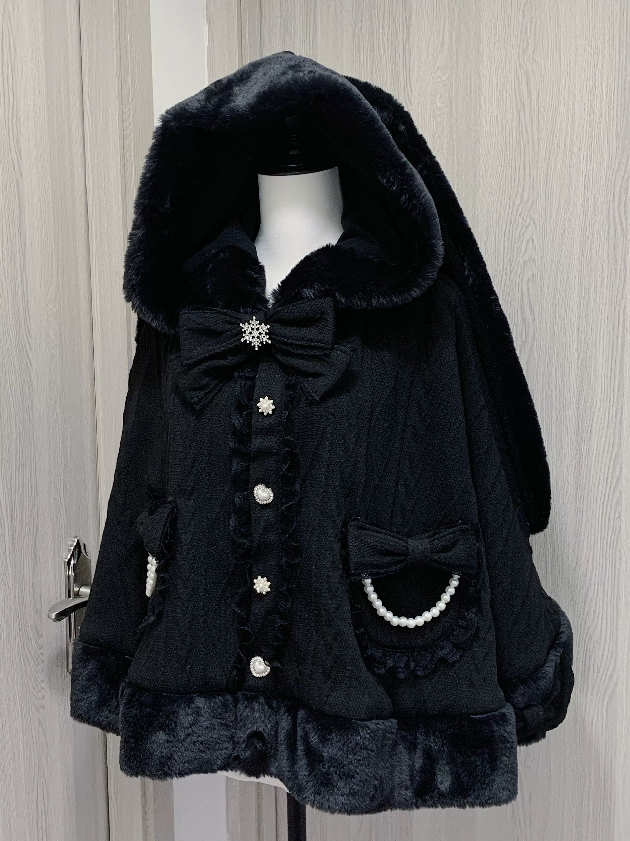 Flat lay product photography of black rabbit ear fleece hooded cape coat with detachable bow tie, showing full garment construction and button closure details