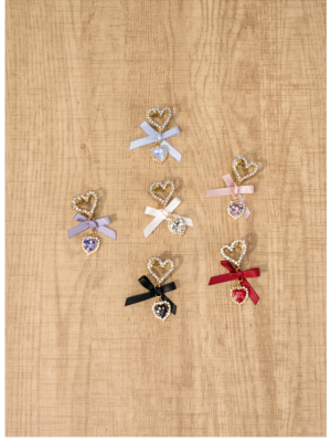 Top-down flat lay of six jirai kei heart-shaped rhinestone hair clips in black dusty pink white light blue purple and red colors with delicate satin bows on wooden texture background