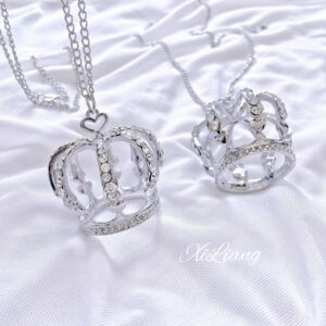 Crown rhinestone necklace with heart-shaped clasp on white satin background, silver-toned hollow crown pendant with sparkling rhinestones, flat lay jewelry photography