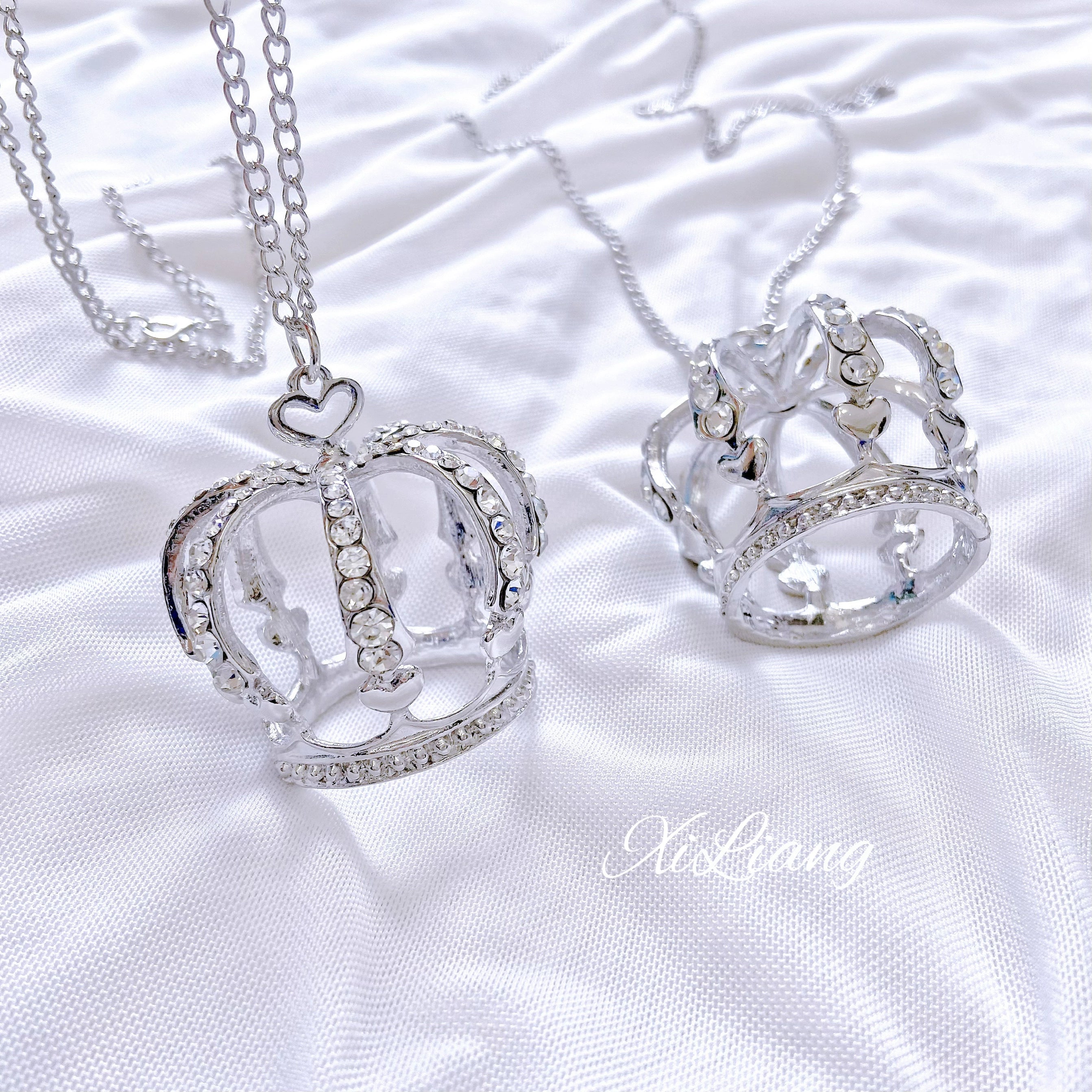 Crown rhinestone necklace with heart-shaped clasp on white satin background, silver-toned hollow crown pendant with sparkling rhinestones, flat lay jewelry photography