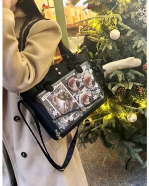 Black Jirai Kei ita bag with clear anime badge display window and heart-shaped metal studs, worn crossbody with side-carrying view next to festive Christmas decorations, showing wavy edge design and bow-adorned shoulder strap
