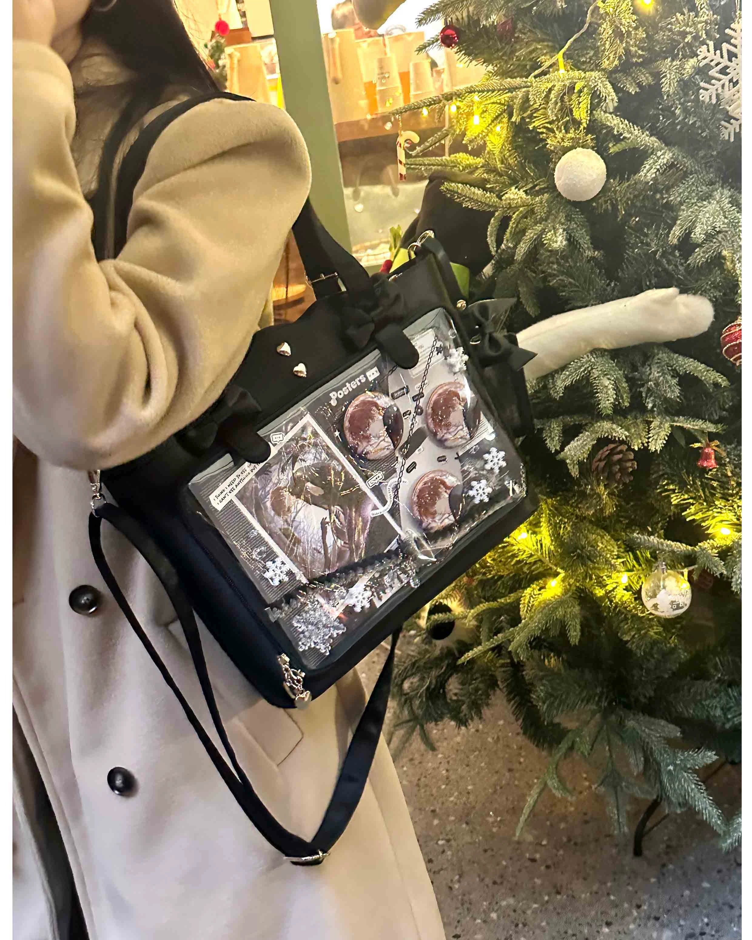 Black Jirai Kei ita bag with clear anime badge display window and heart-shaped metal studs, worn crossbody with side-carrying view next to festive Christmas decorations, showing wavy edge design and bow-adorned shoulder strap