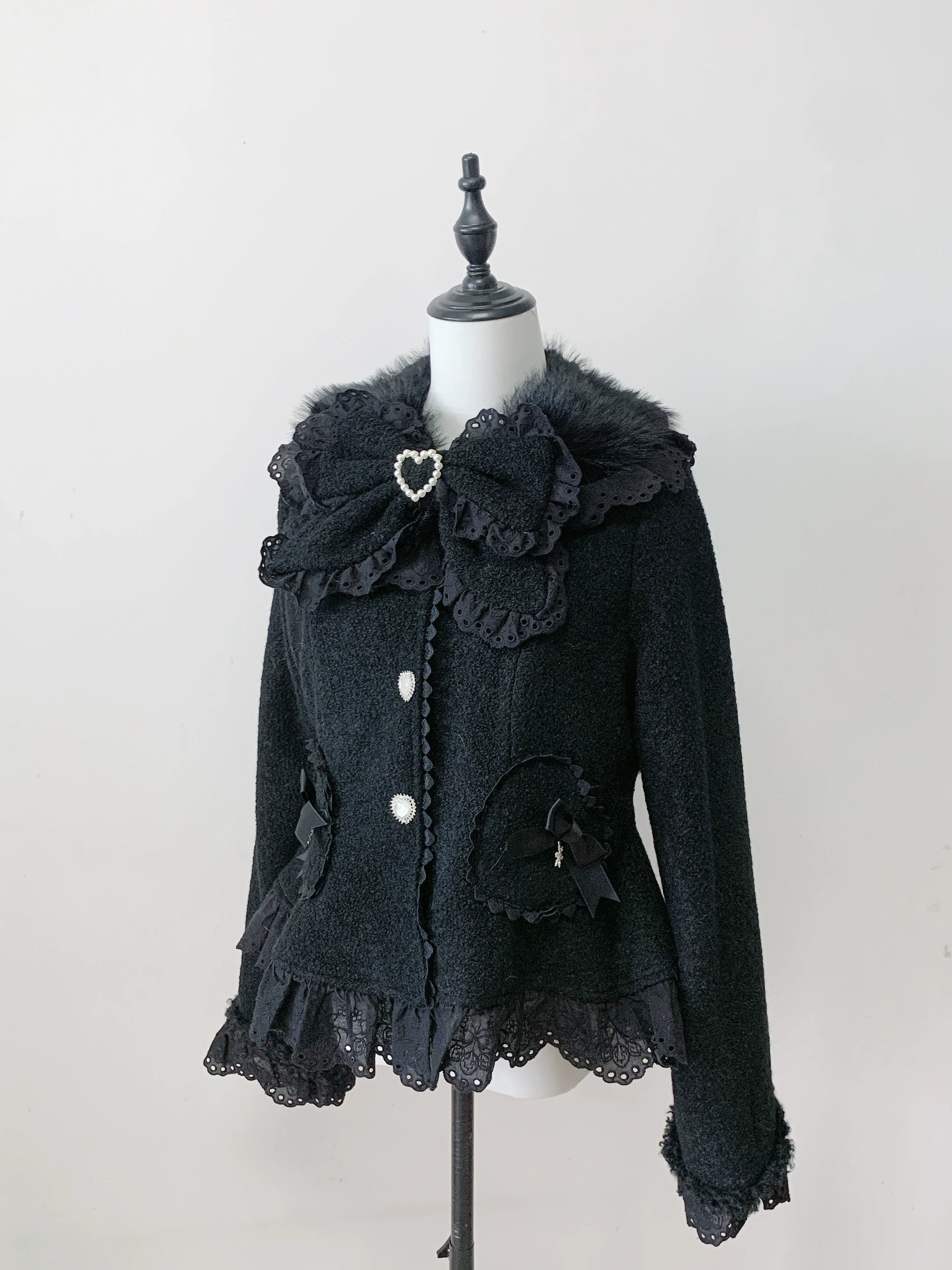 Black Ryousangata Jirai Kei wool coat with heart brooch and ruffled hem, mannequin studio display showing single-breasted button closure and large bow collar