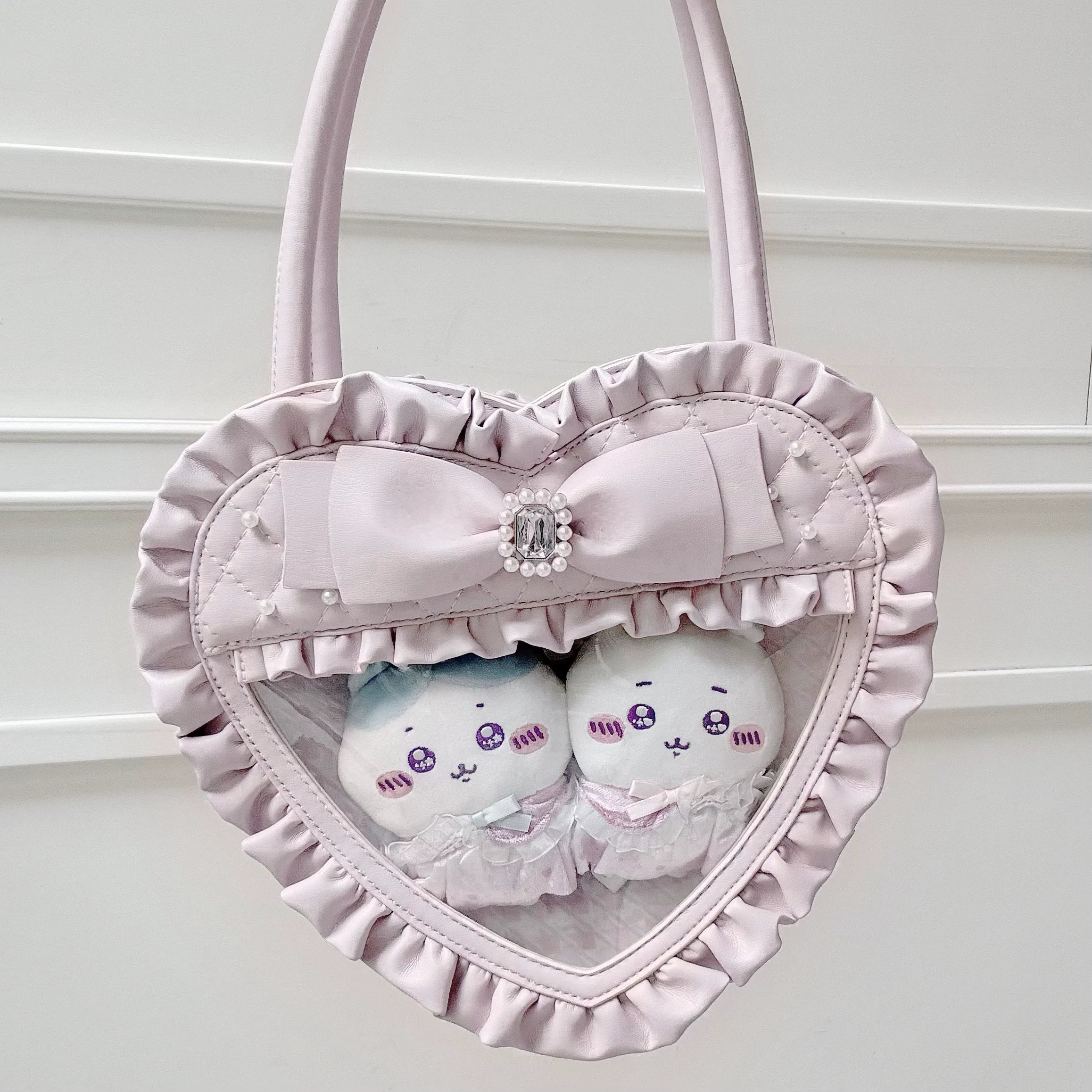 Light pink heart-shaped Jirai Kei itabag close-up front view against white wall featuring clear acrylic window with white plush dolls, ruffled edge and rhinestone decorated bow