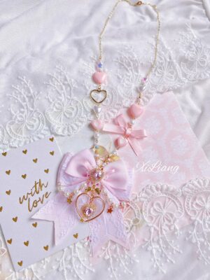 Long pink Lolita sweater necklace featuring hollow gold heart pendant, pearl beads, and decorative pink bow detail arranged on lace cloth with greeting card, overhead flat lay